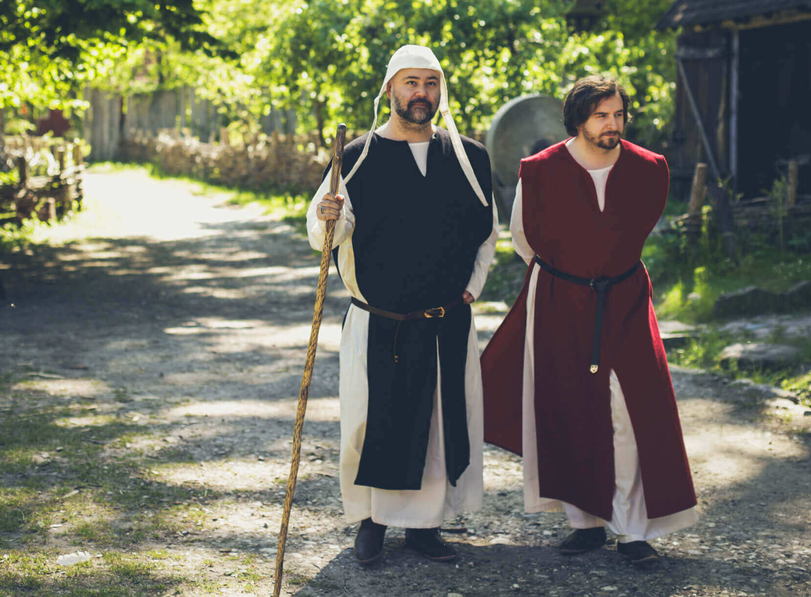 Two men dressed in medieval costumes walking along a rustic path in a sunny outdoor setting.
