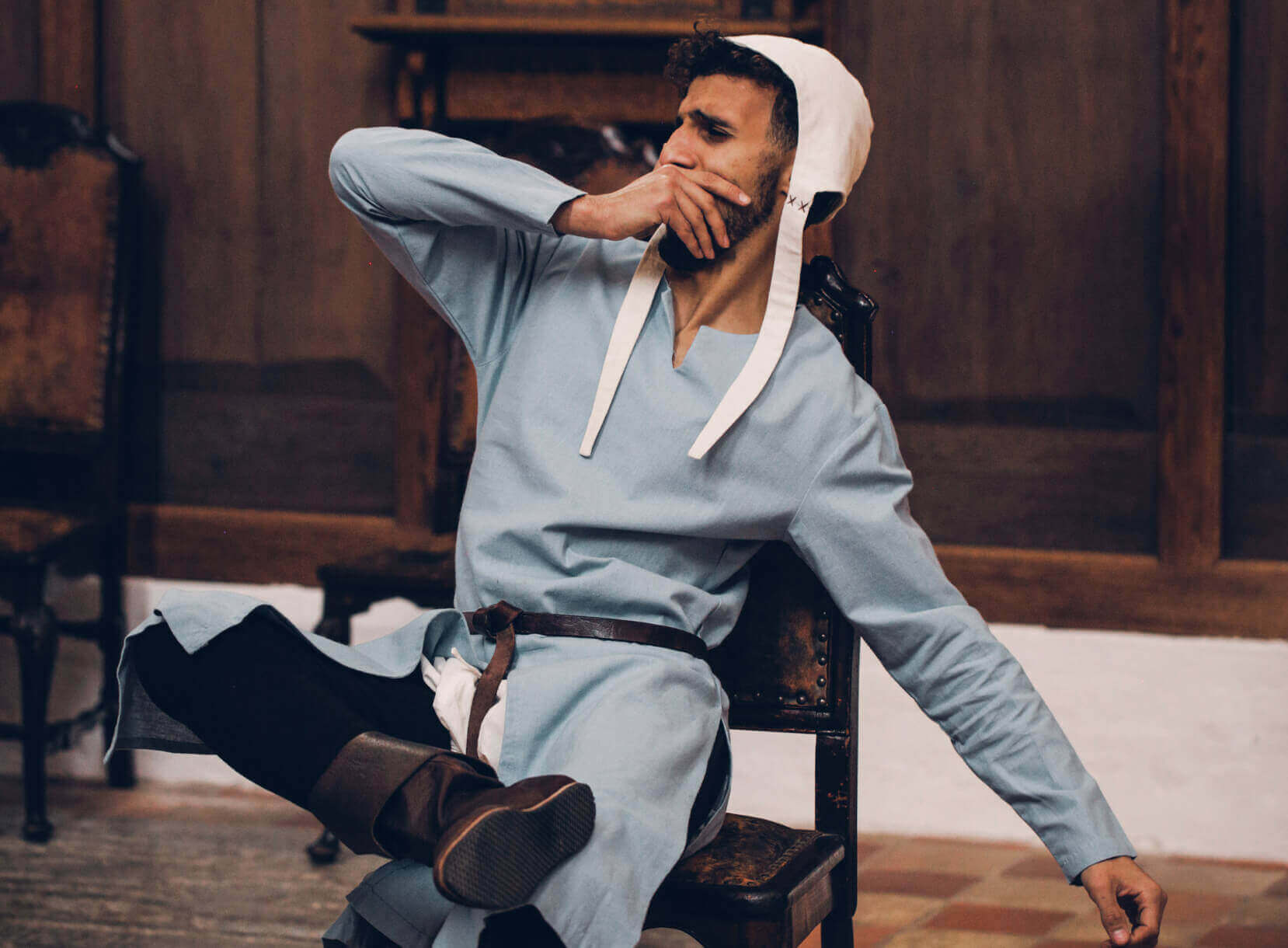 Man in light blue tunic, white coif, and brown boots sitting in high medieval attire.