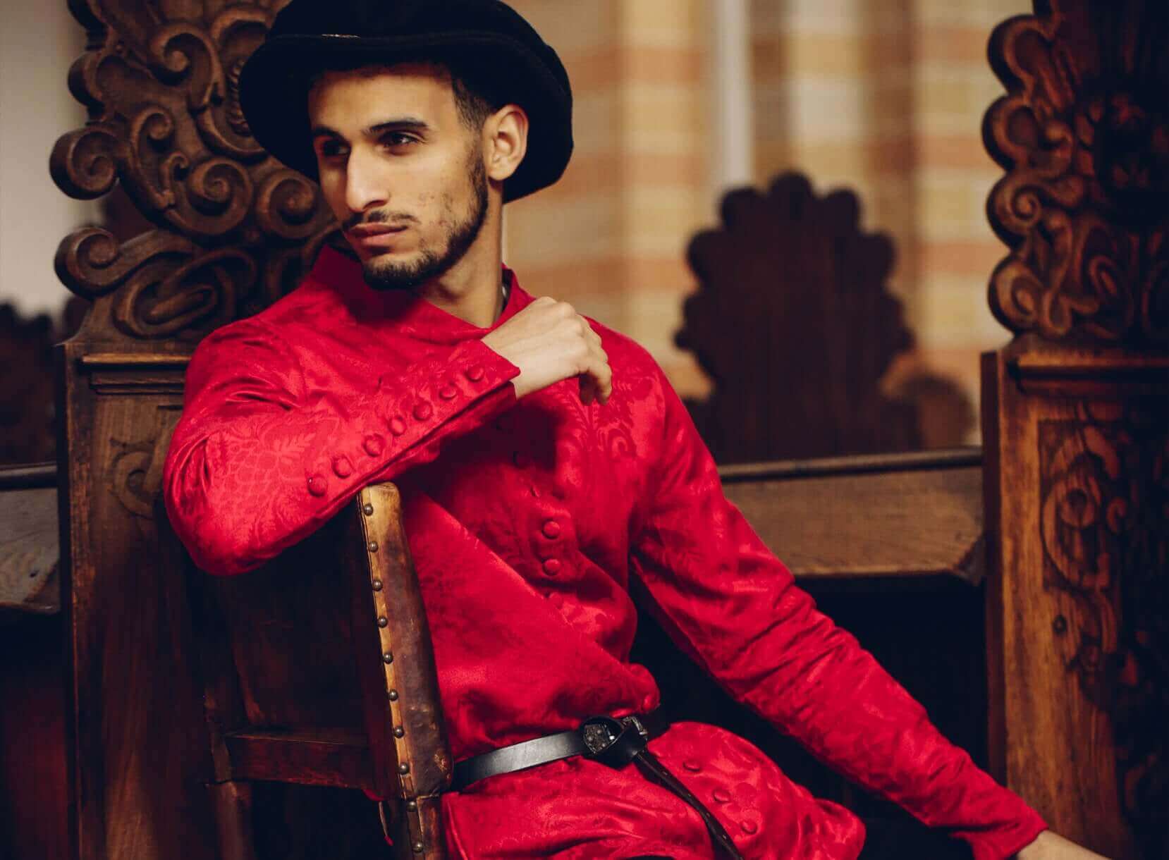 Man in red medieval tunic and black hat sitting on a carved wooden chair.