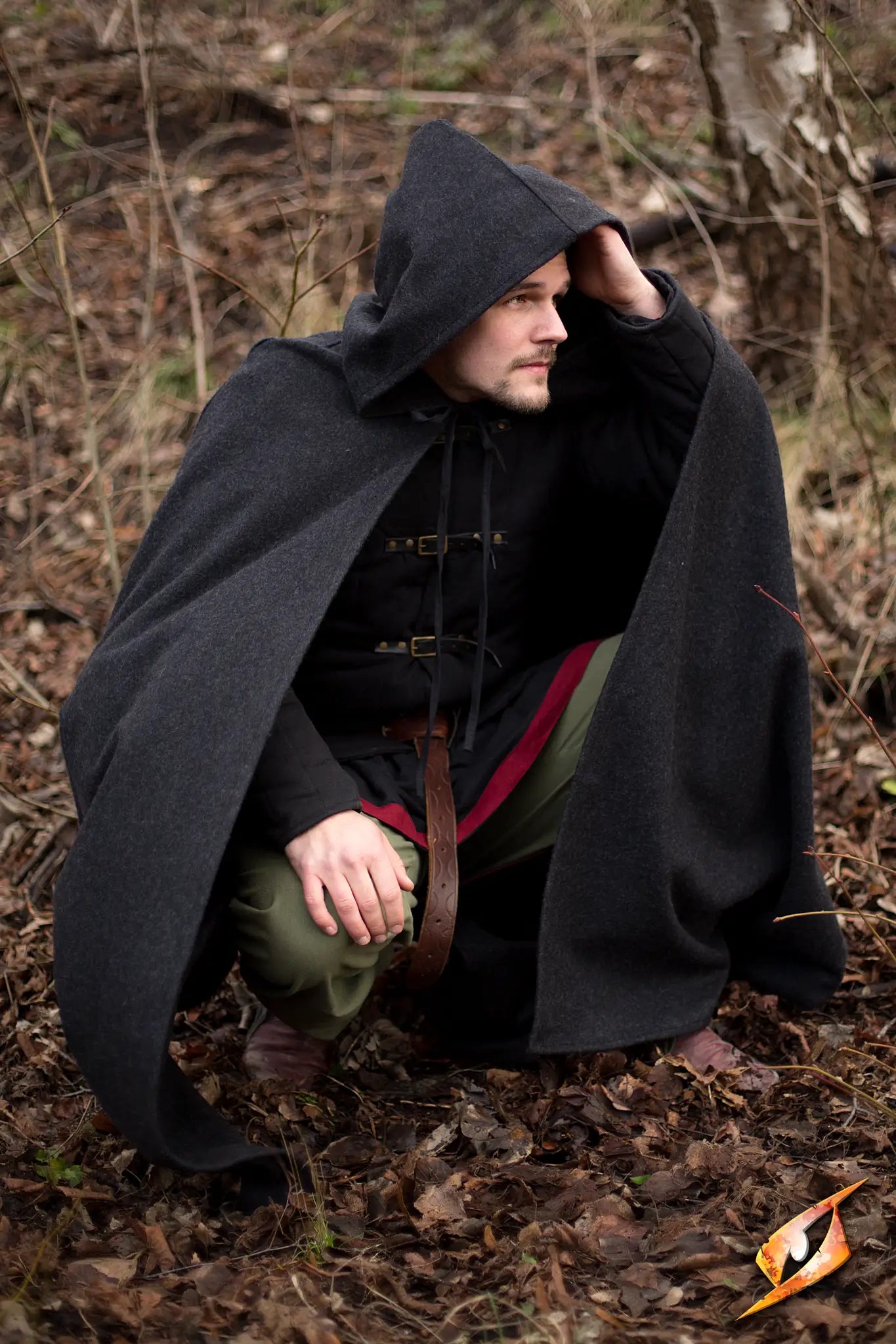 Man wearing Cape Wool cape, crouching in nature, featuring deep hood and form-fitting shoulders.