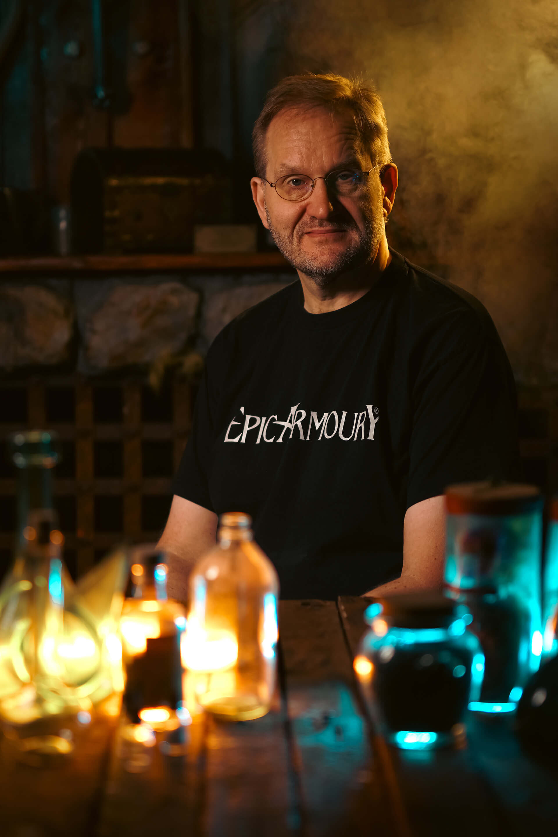 Man in a black shirt sitting at a table with glowing bottles, creating a mystical atmosphere.