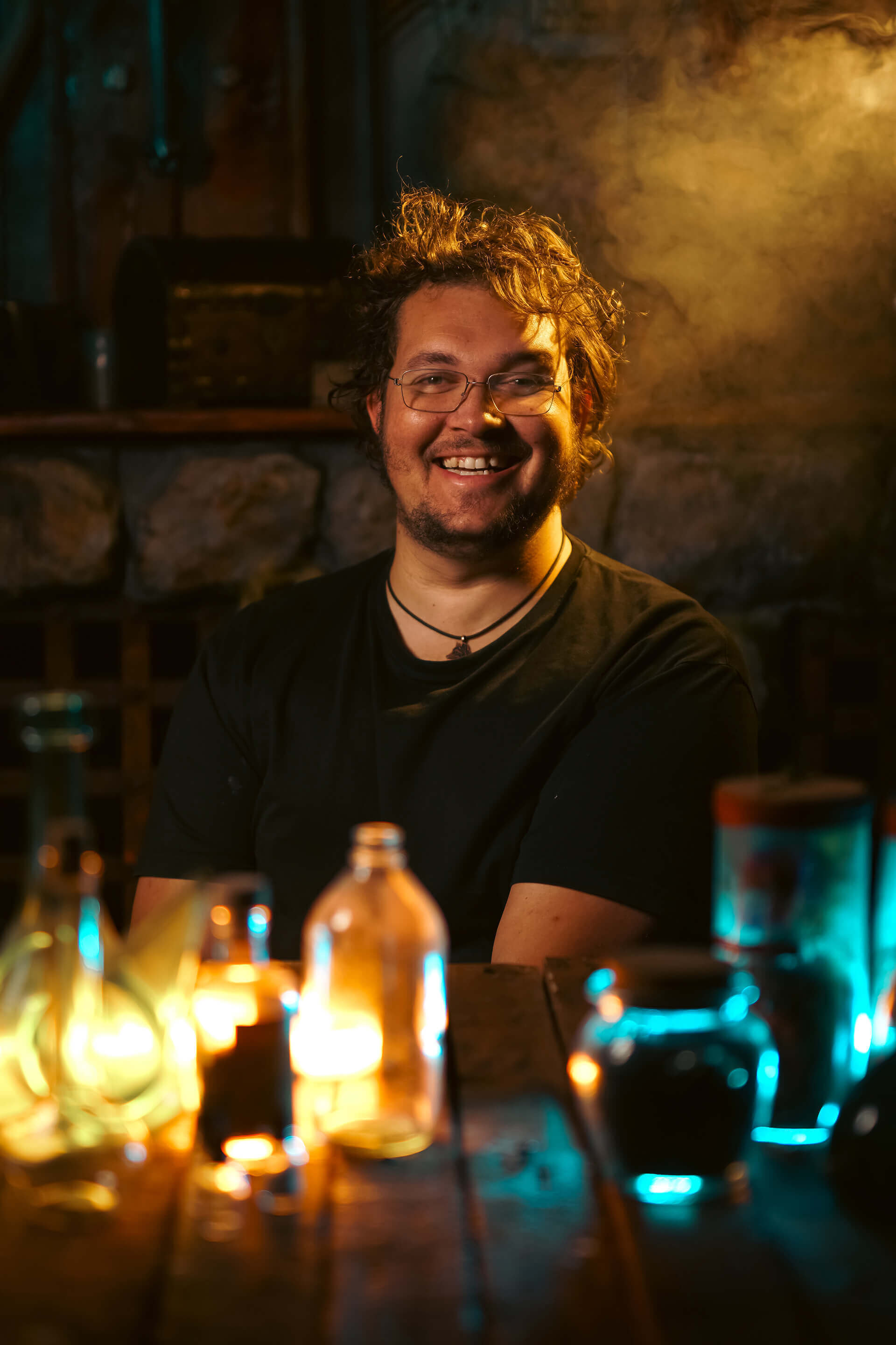 Smiling man in a dimly lit setting with colorful potion bottles on a wooden table.