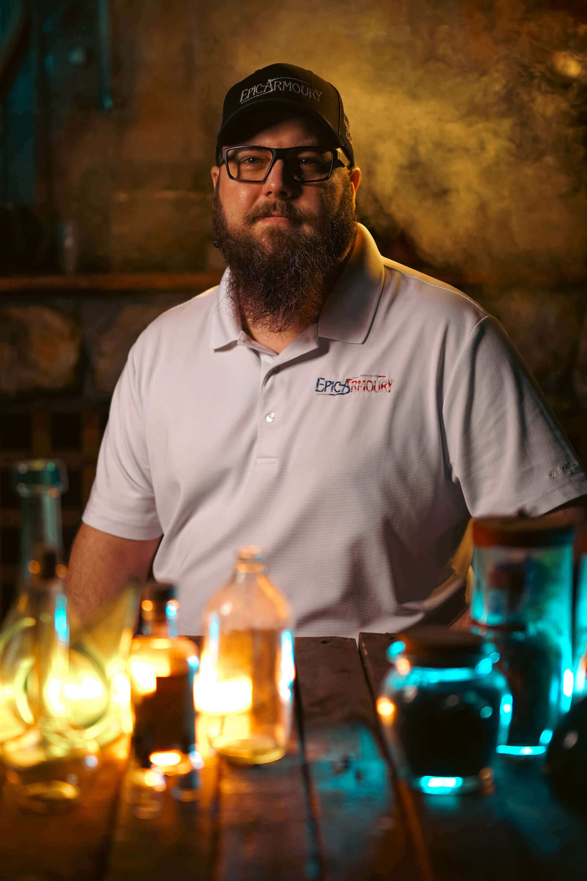Bearded man in a white polo shirt posing with colorful glowing liquids in a science-themed setting.