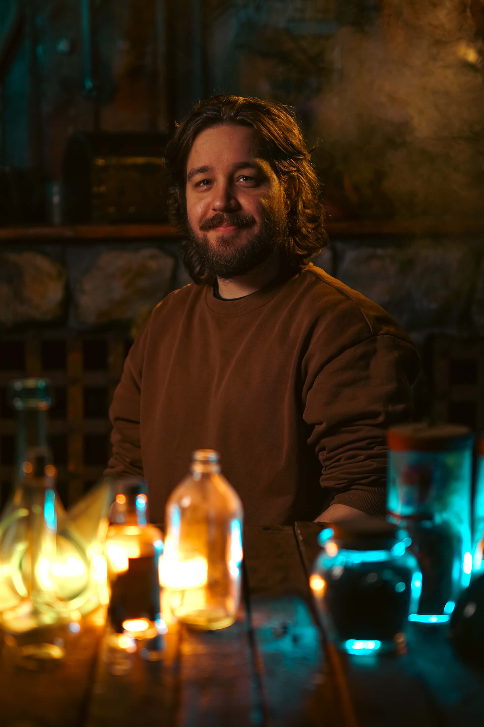 Smiling man in a cozy ambient setting surrounded by colorful glass bottles and potions on a wooden table.