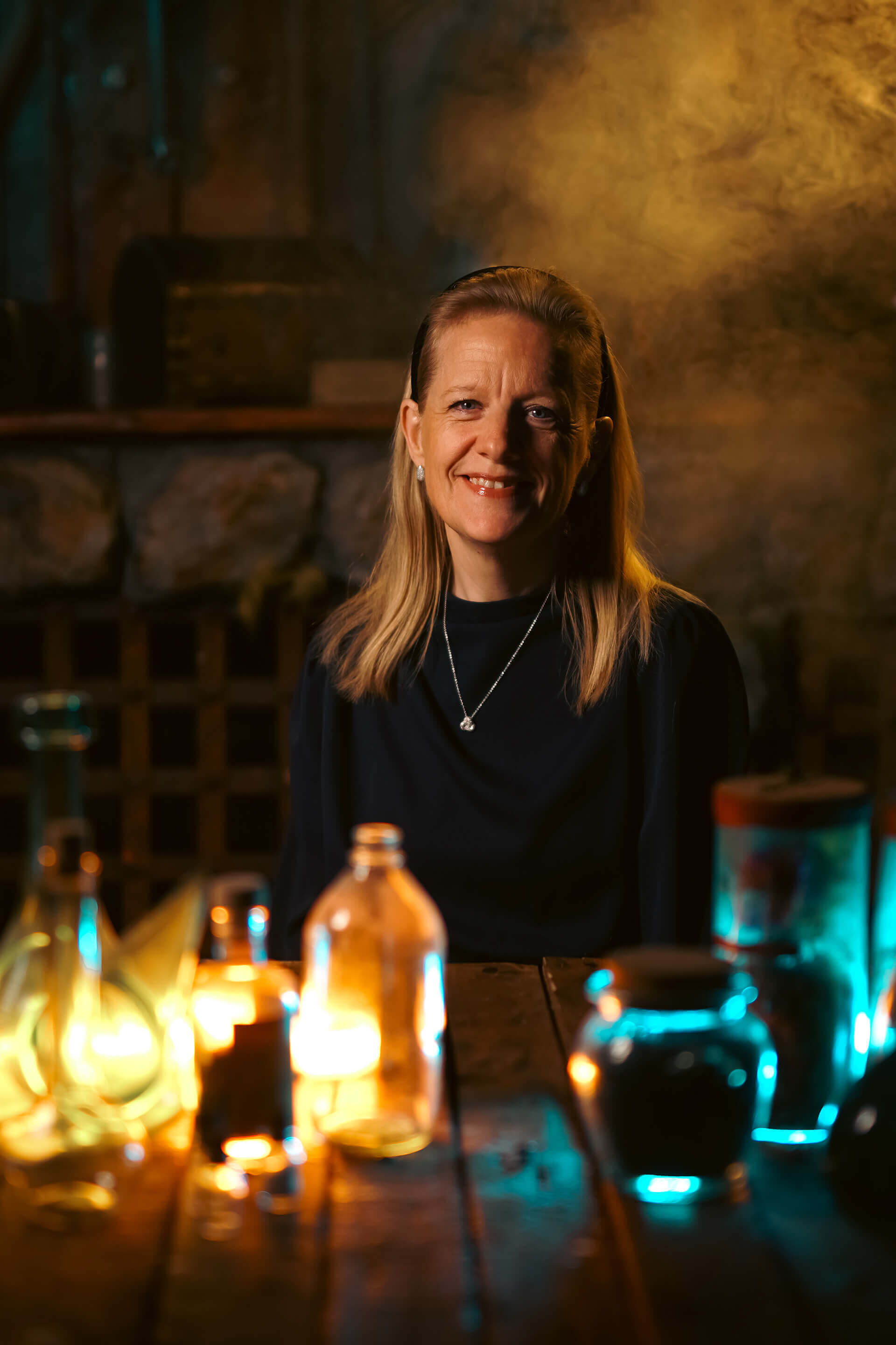 A woman with long blonde hair smiling in a dimly lit room surrounded by glowing bottles and smoke.