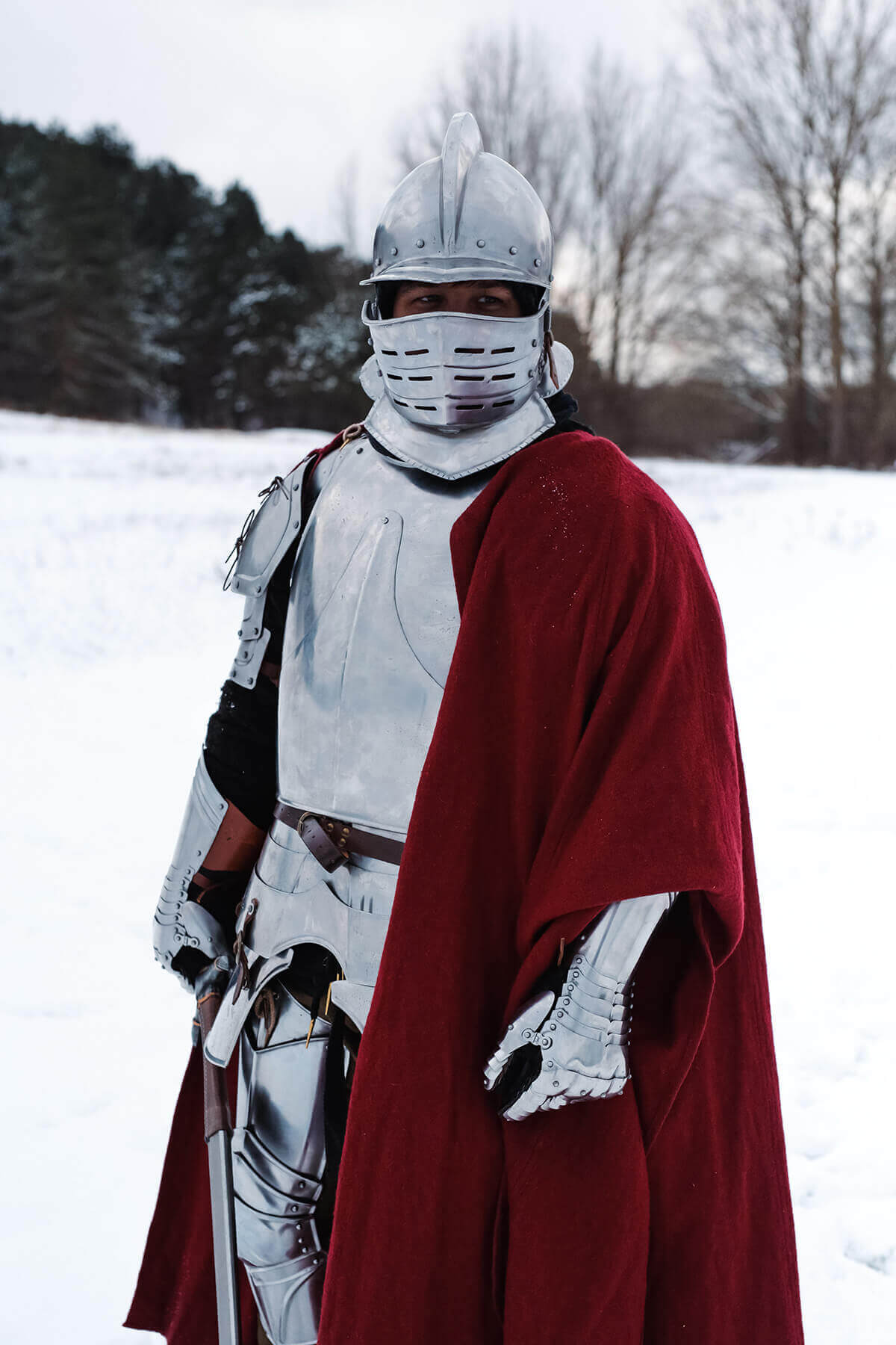 Knight in medieval armor with a red cape standing in a snowy landscape, showcasing historical attire.