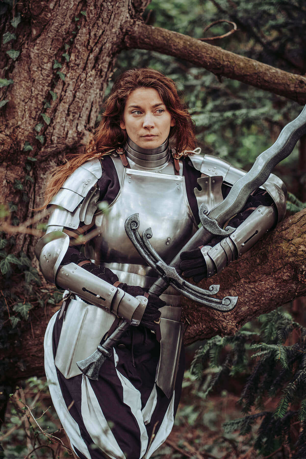 A woman in medieval armor resting against a tree, holding a sword, exuding strength and bravery.