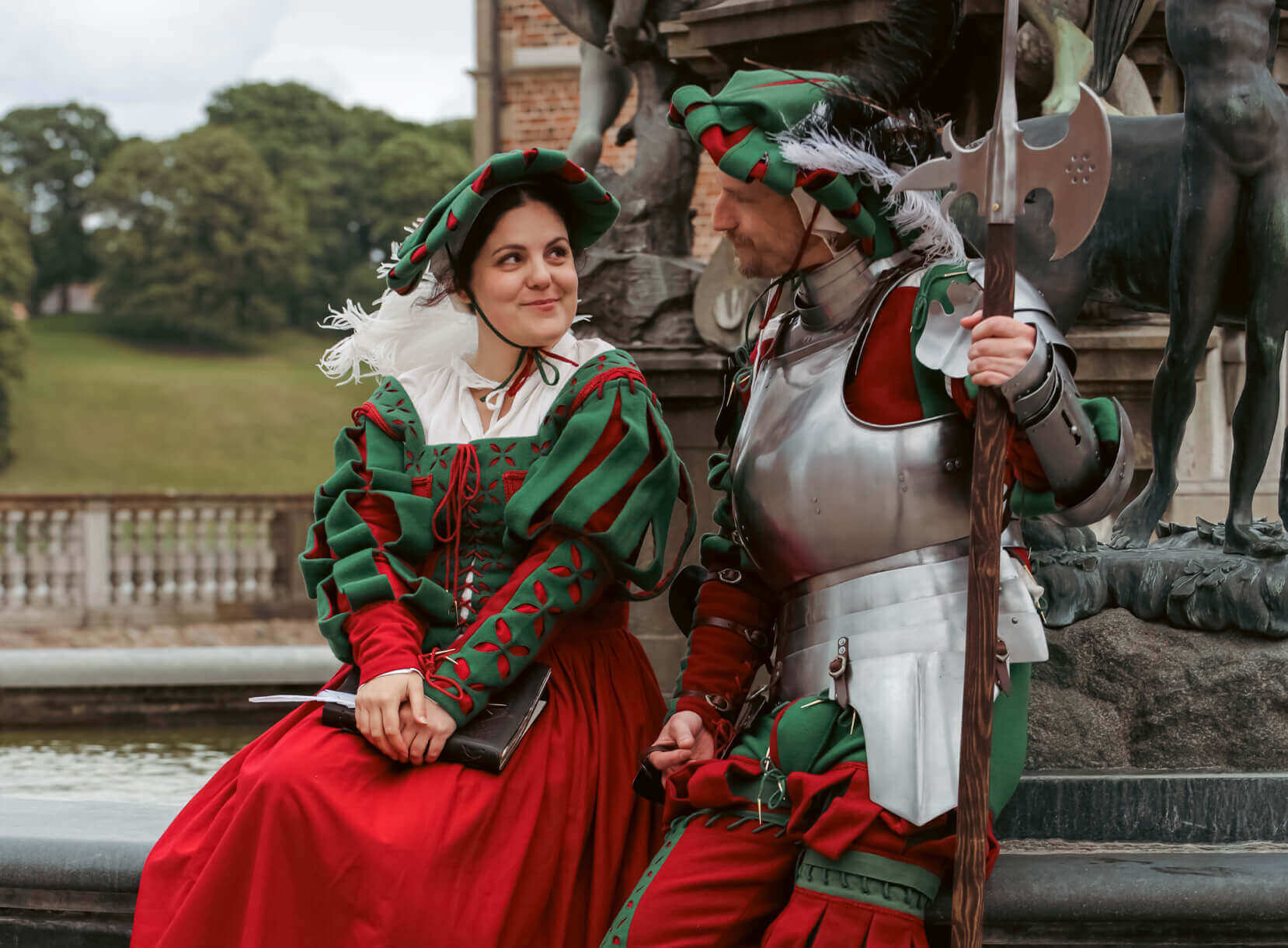 A woman in a red and green dress and a man in silver armor engage in conversation near a fountain.