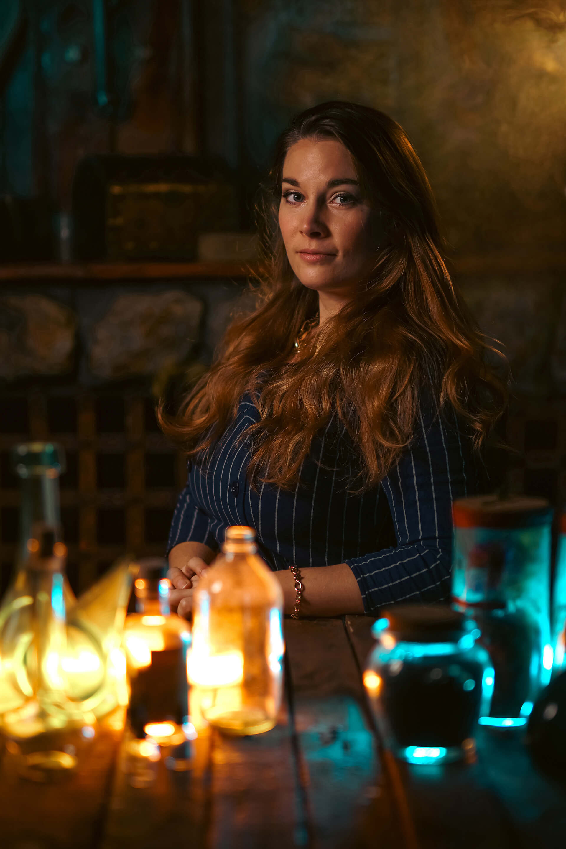 Portrait of a woman with long hair, seated at a table with colorful glowing bottles and a moody atmosphere.