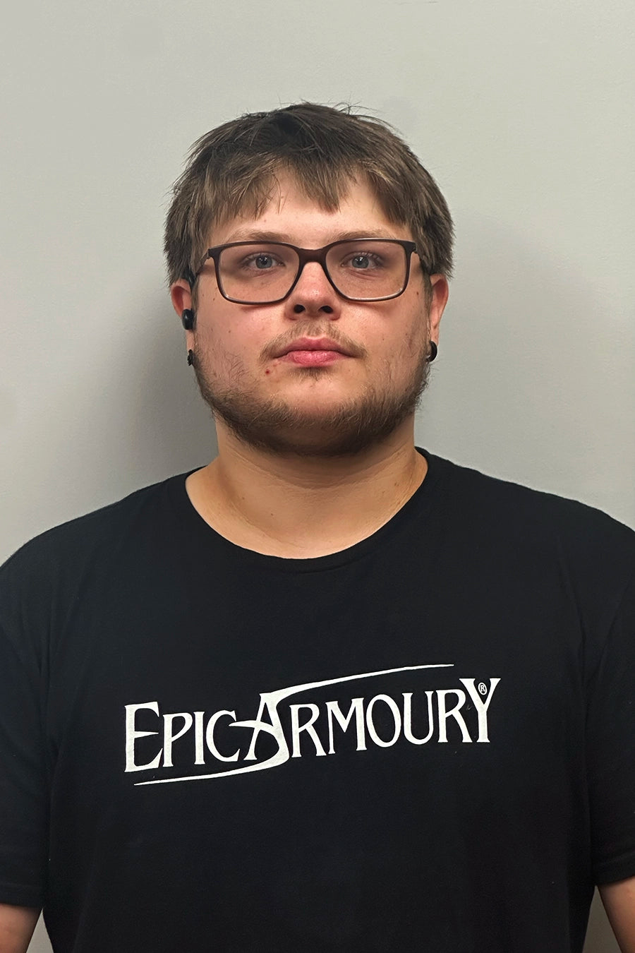 Portrait of a young man wearing glasses and a black Epic Armoury t-shirt, against a plain background.