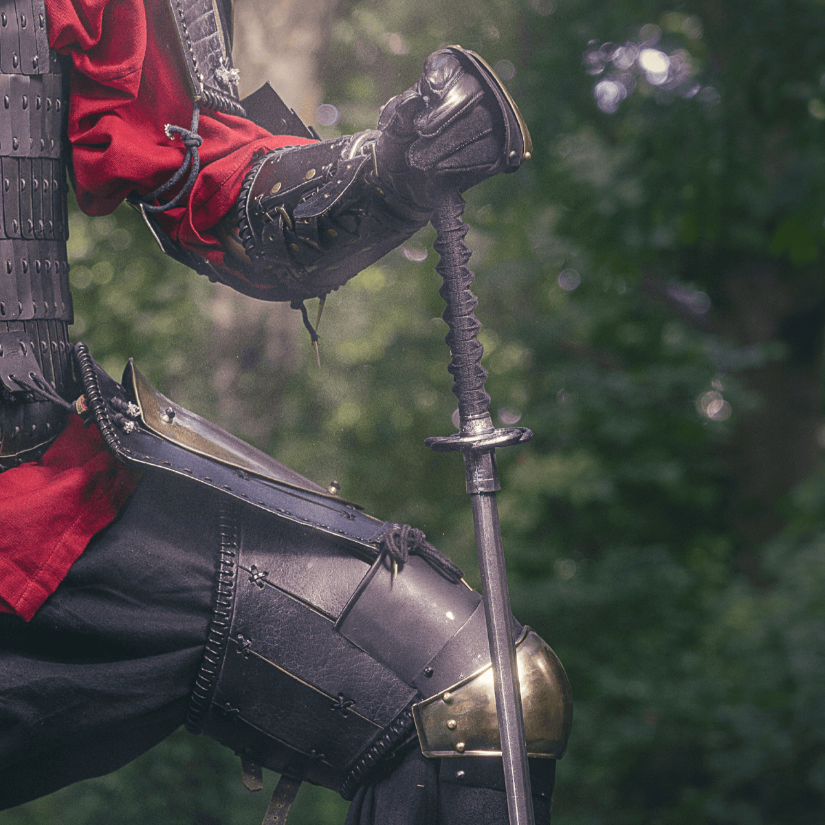Close-up of a medieval knight in armor holding a sword, dressed in red and black, standing in a forest backdrop.