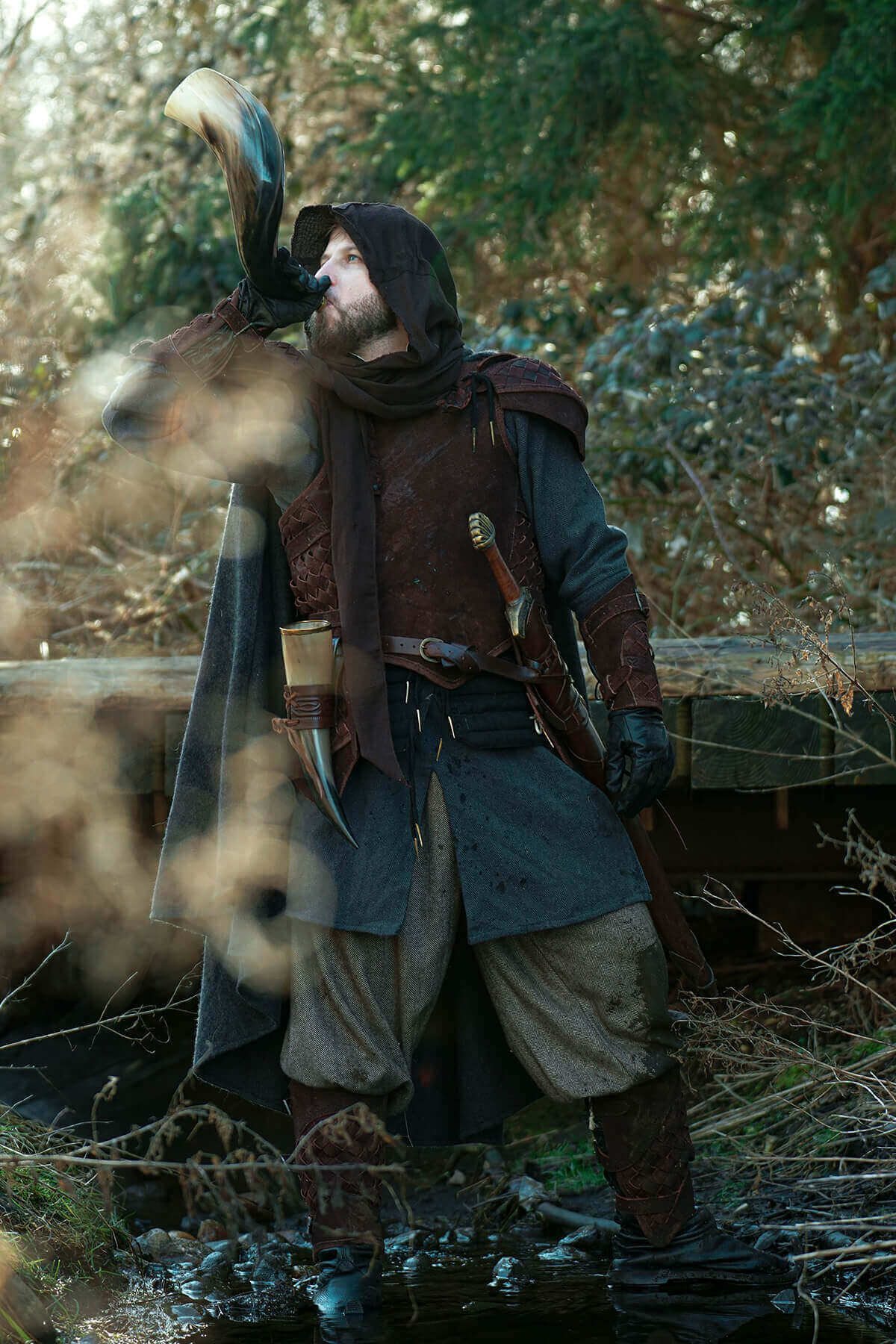 A cloaked figure in a forest drinking from a horn, showcasing detailed costume and armaments.