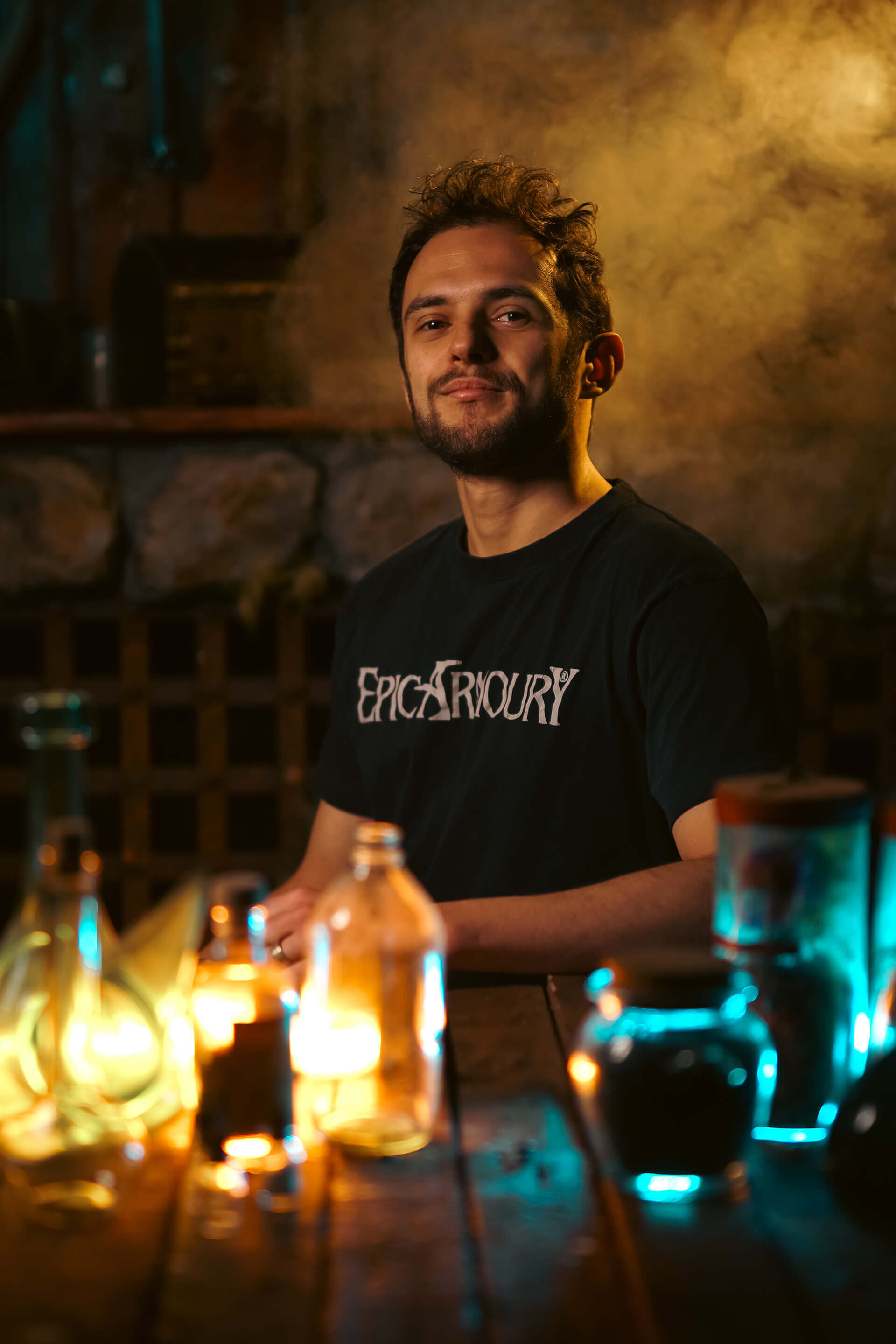 Smiling man wearing an Epic Armoury shirt, surrounded by glowing potion bottles in a cozy setting.