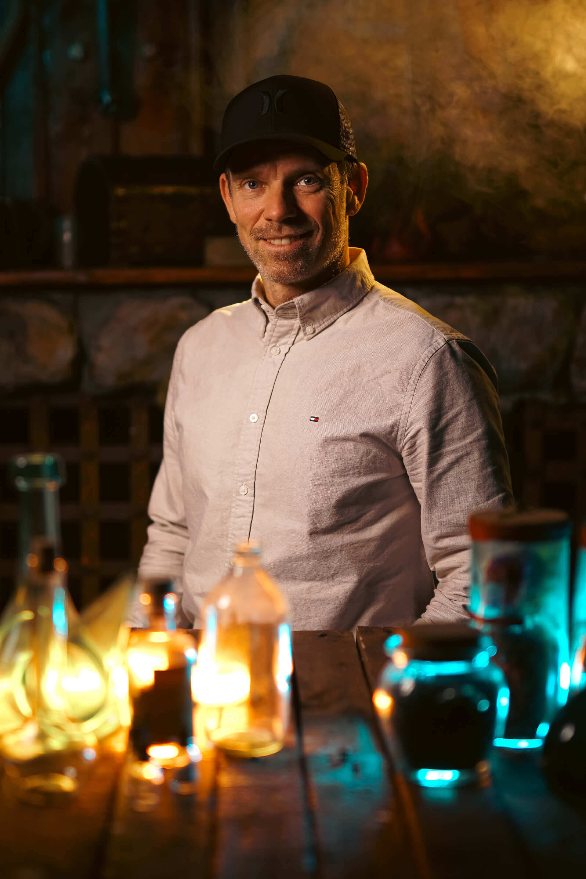 Man with a cap sitting at a table with glowing bottles in a dimly lit atmosphere.