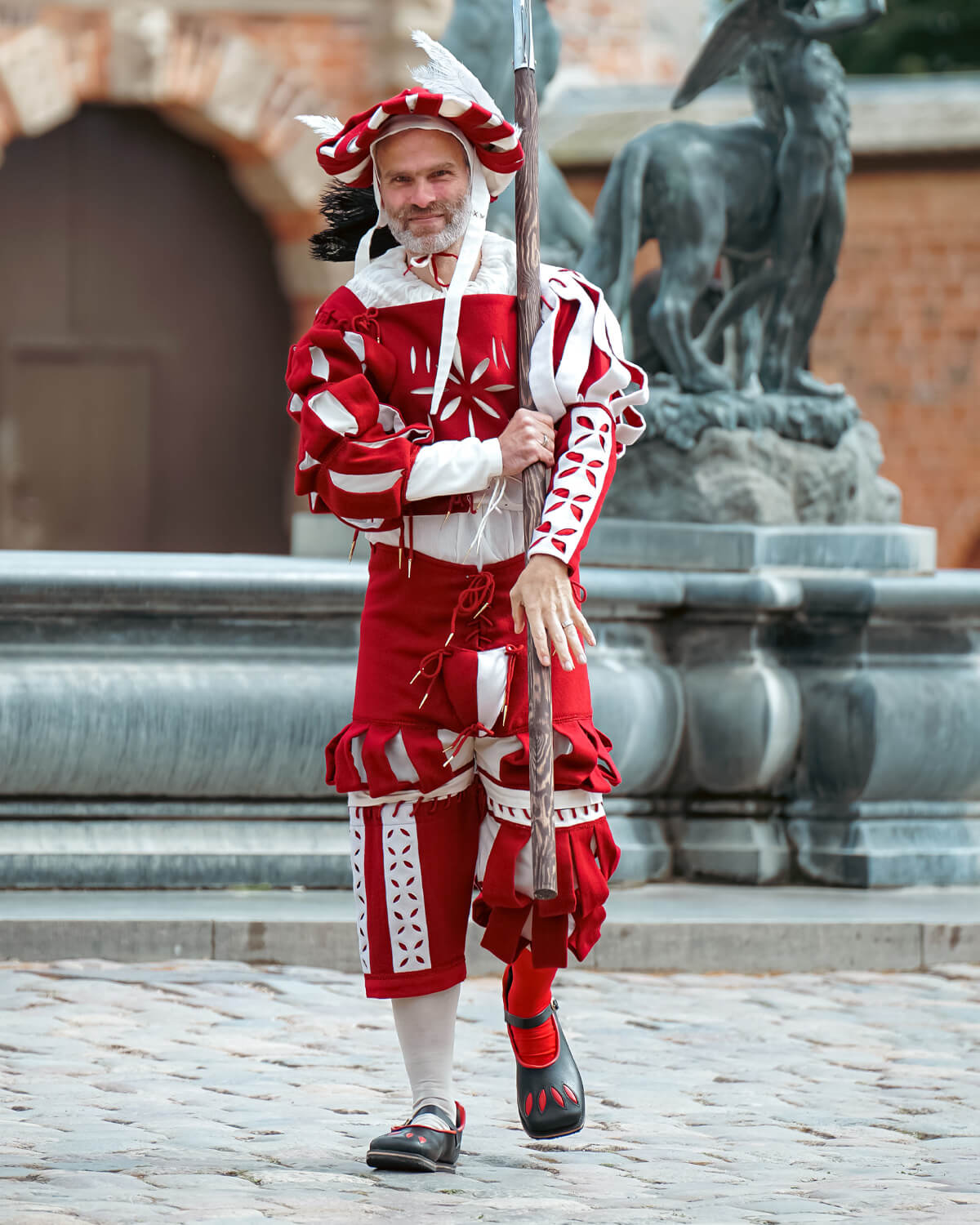 Man in colorful red and white traditional costume carrying a spear, standing near a fountain.