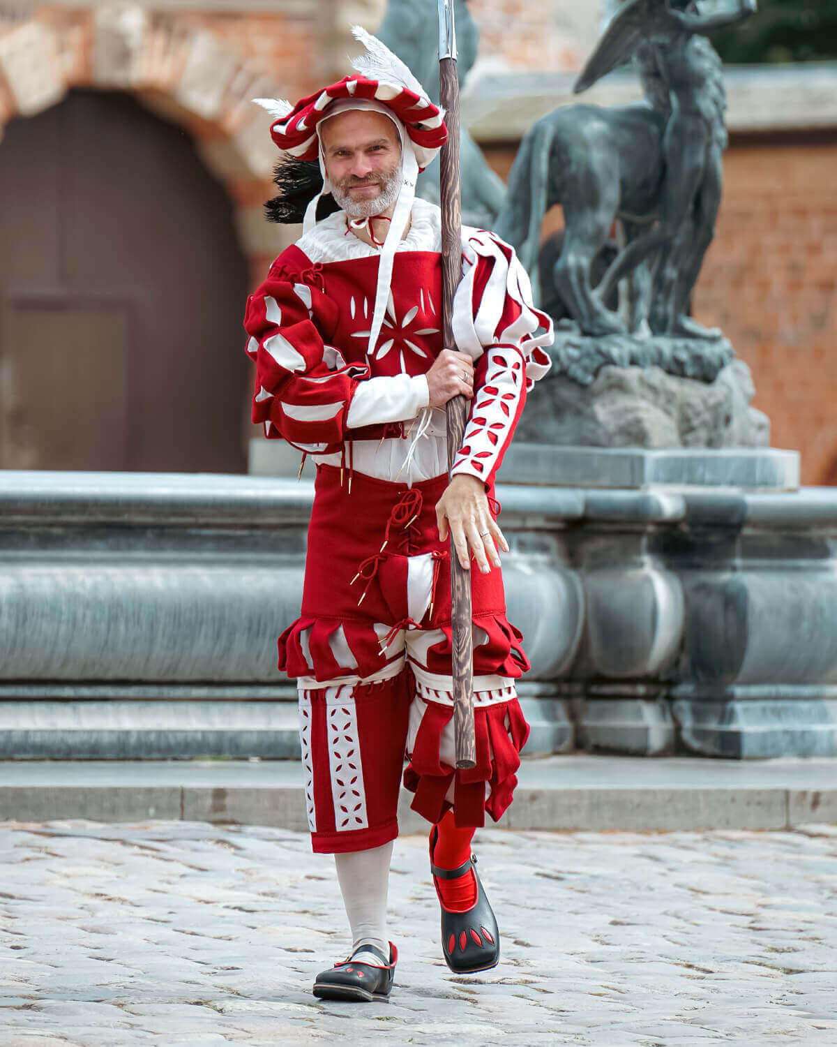 Man in colorful red and white traditional costume carrying a spear, standing near a fountain.