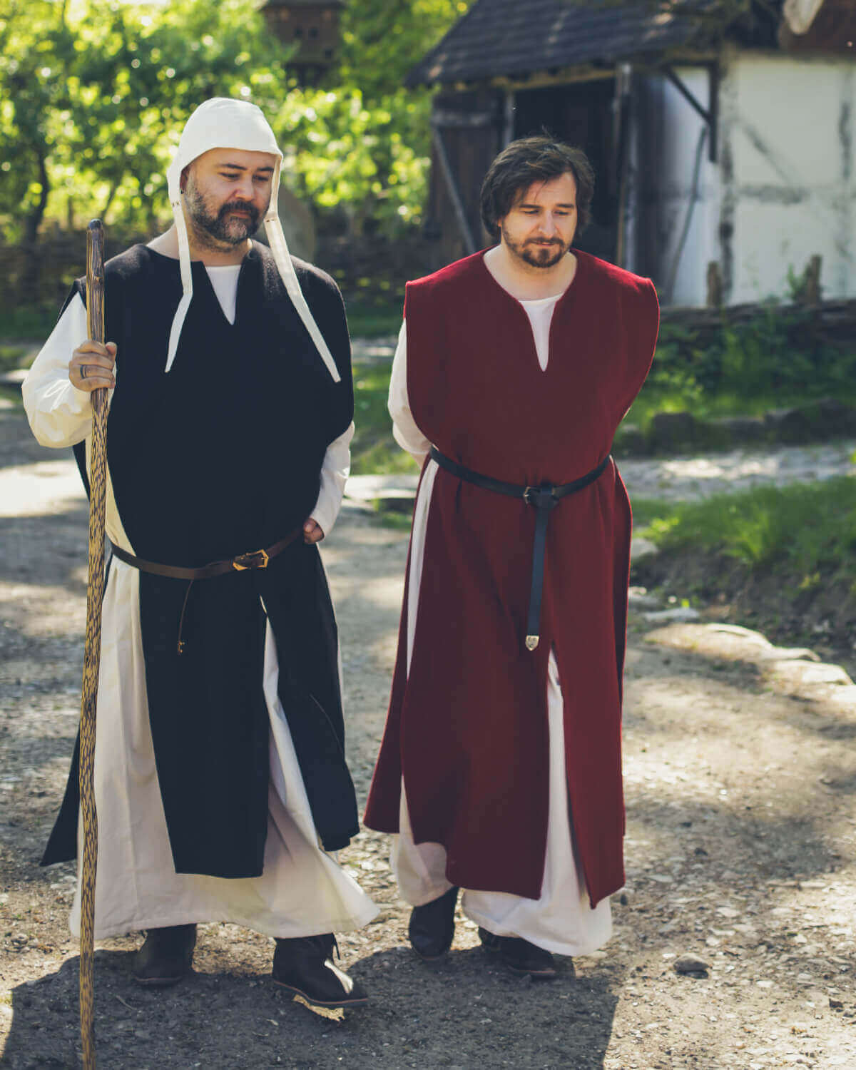 Two men in medieval attire, one in black and one in red, walking together along a rustic path.