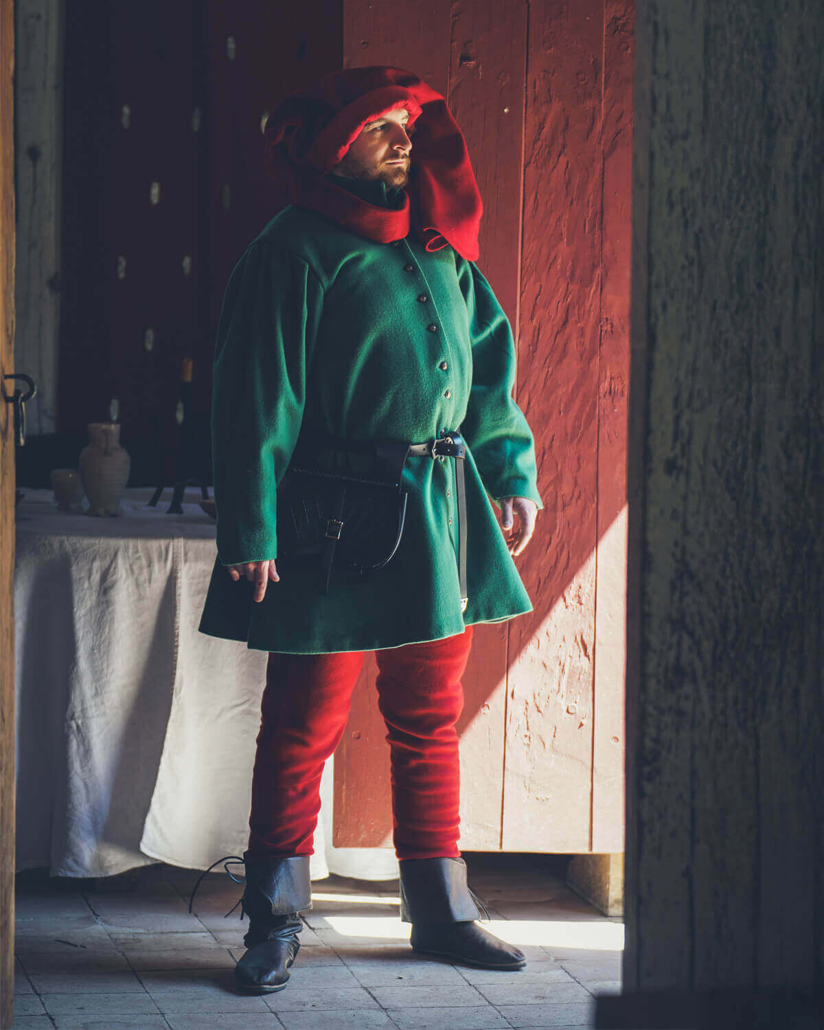 Man dressed in historical attire with a green coat and red pants in a rustic setting.