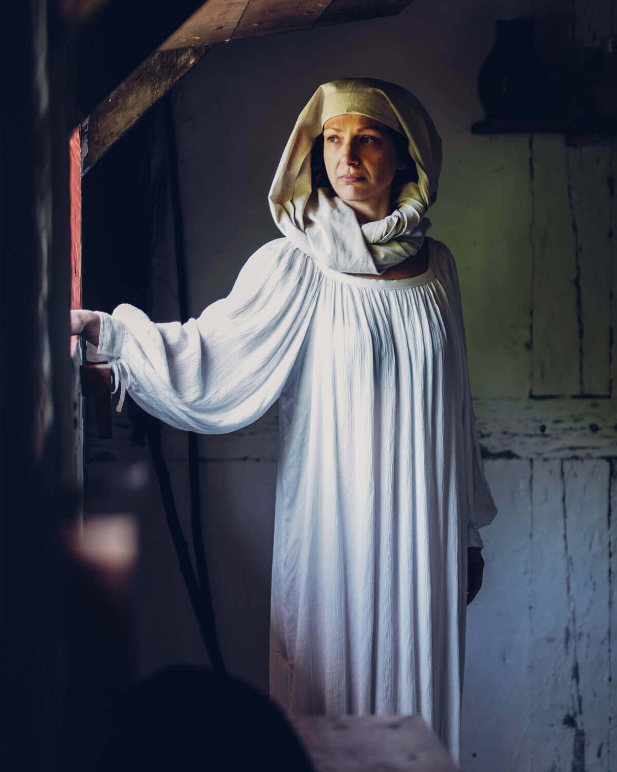Woman in historical white dress and headscarf, standing by window, gazing thoughtfully in a rustic setting.
