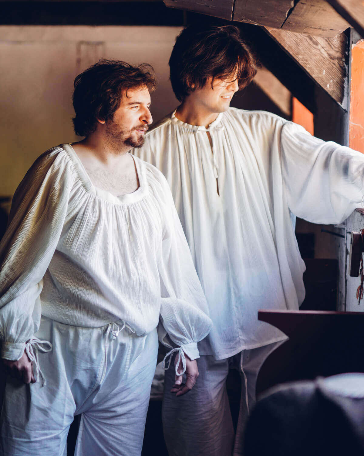 Two men in white vintage clothing, standing by a window, showcasing a historical theme and relaxed atmosphere.
