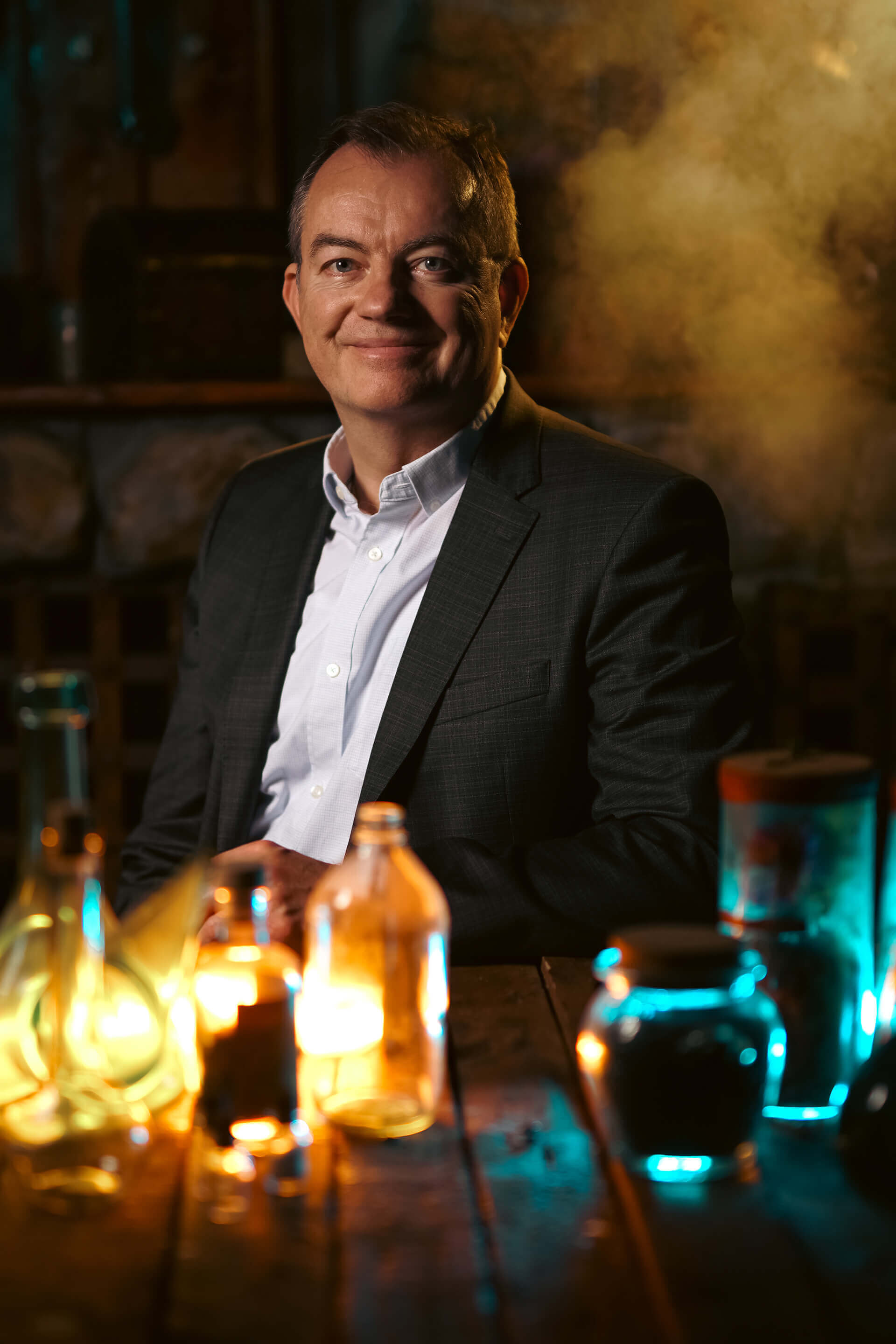 Man in a suit smiling against a backdrop of colorful glass bottles and smoke, creating a mysterious ambiance.
