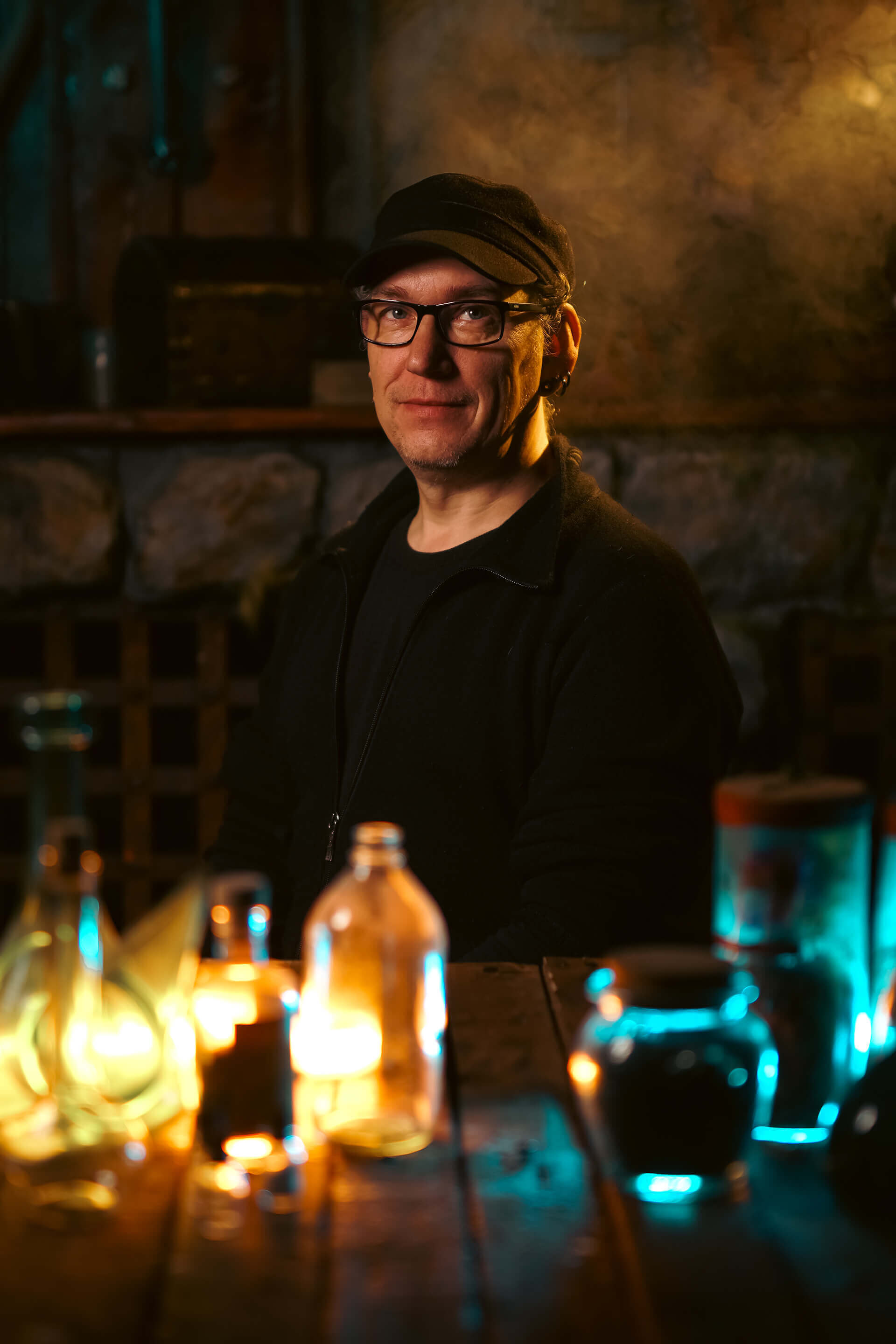 Portrait of a man wearing glasses and a cap, surrounded by glowing bottles in a dimly lit setting.