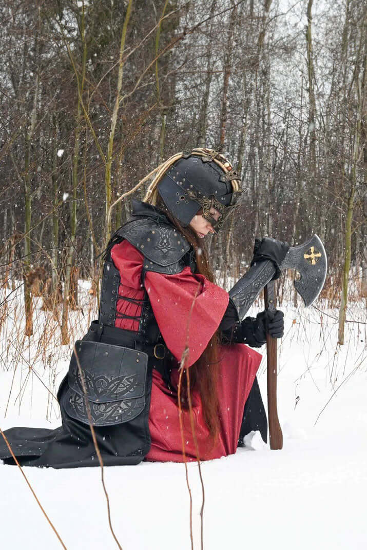 A warrior kneeling in snow, holding the Baruk II axe, showcasing its steel head, gold cross, and light brown handle.