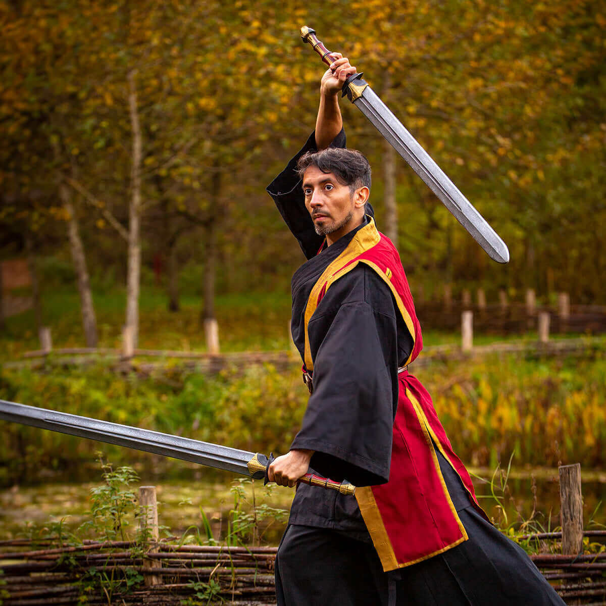 Martial artist in traditional garb wielding dual swords in a wooded outdoor setting.