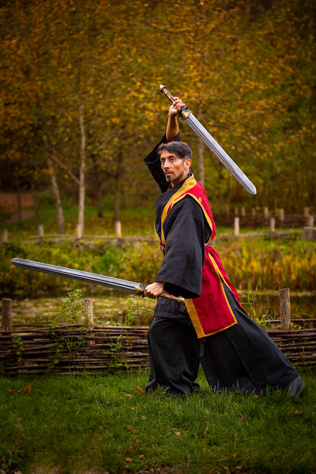Person in historical costume holding two swords outdoors with trees in the background
