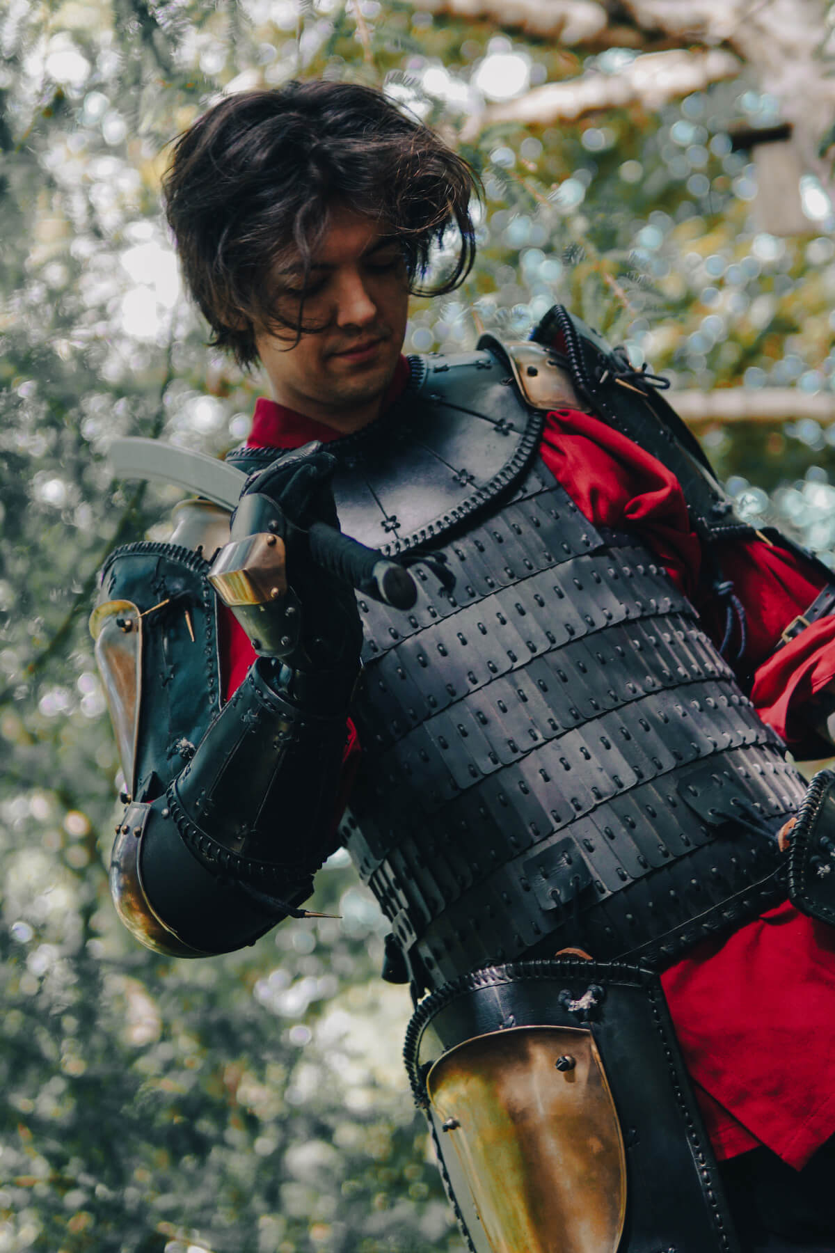 Person wearing black armor with a red shirt, holding a sword outdoors.