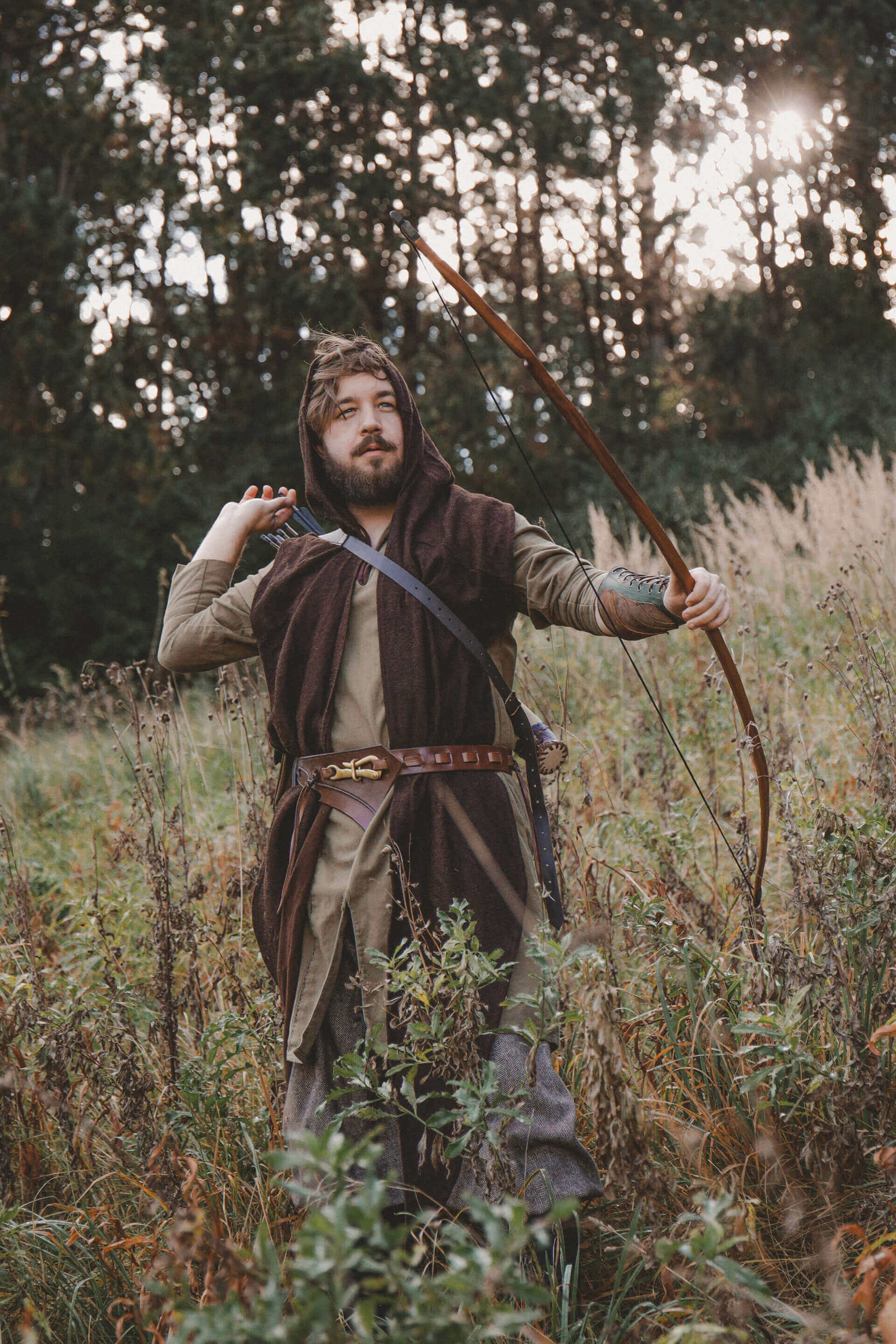 Pretty Man in medieval attire holding a bow and arrow in a forest setting