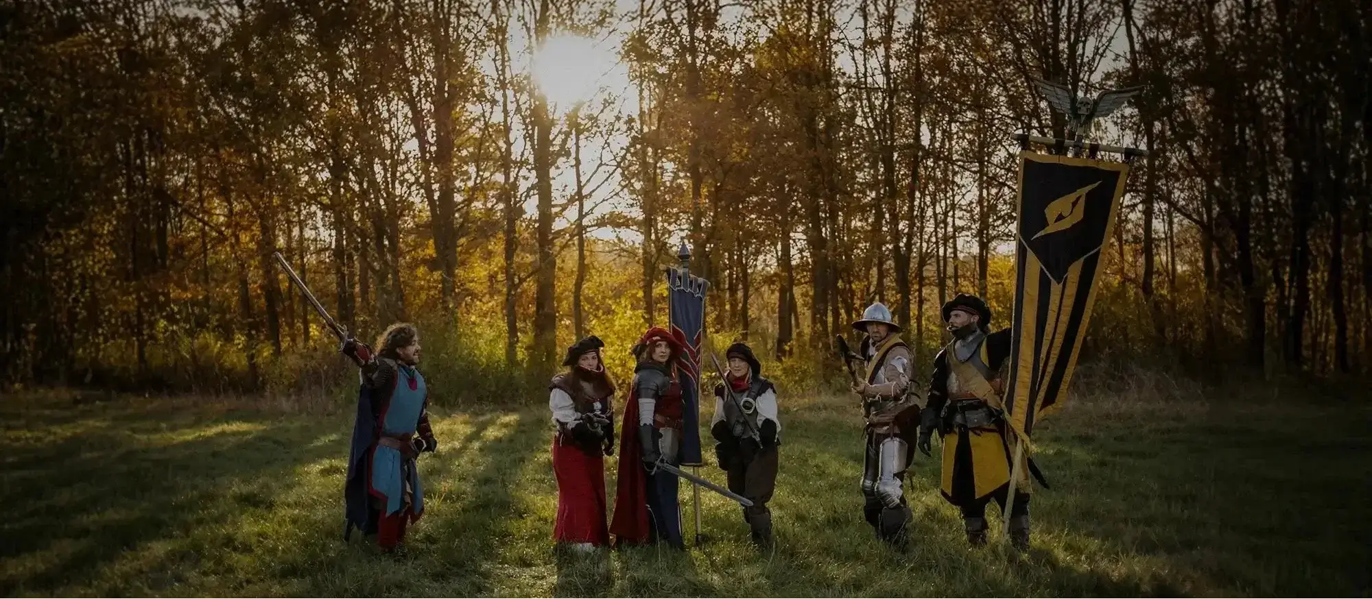 Group of LARP participants in medieval costumes standing in a field at sunset with banners and weapons.