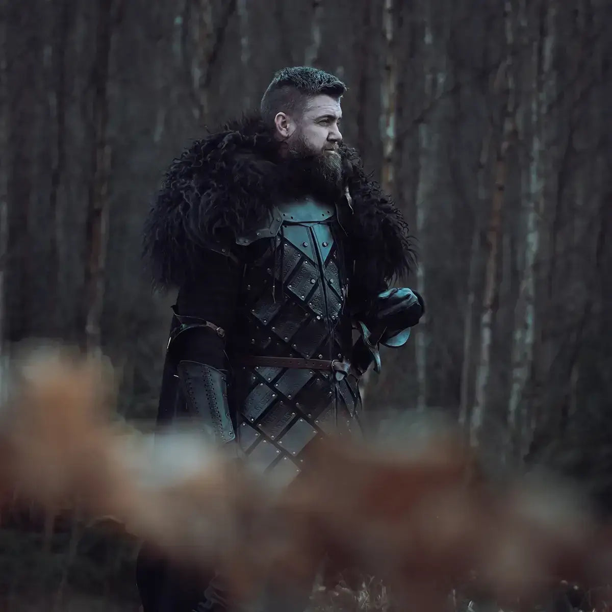 A man dressed in dark medieval armor with a fur collar, standing in a forest setting during dusk.