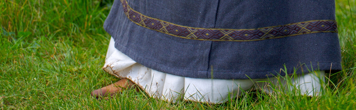 A close-up of a dark gray traditional dress or skirt with a decorative embroidered band, layered over a white underlayer, set against a background of green grass.