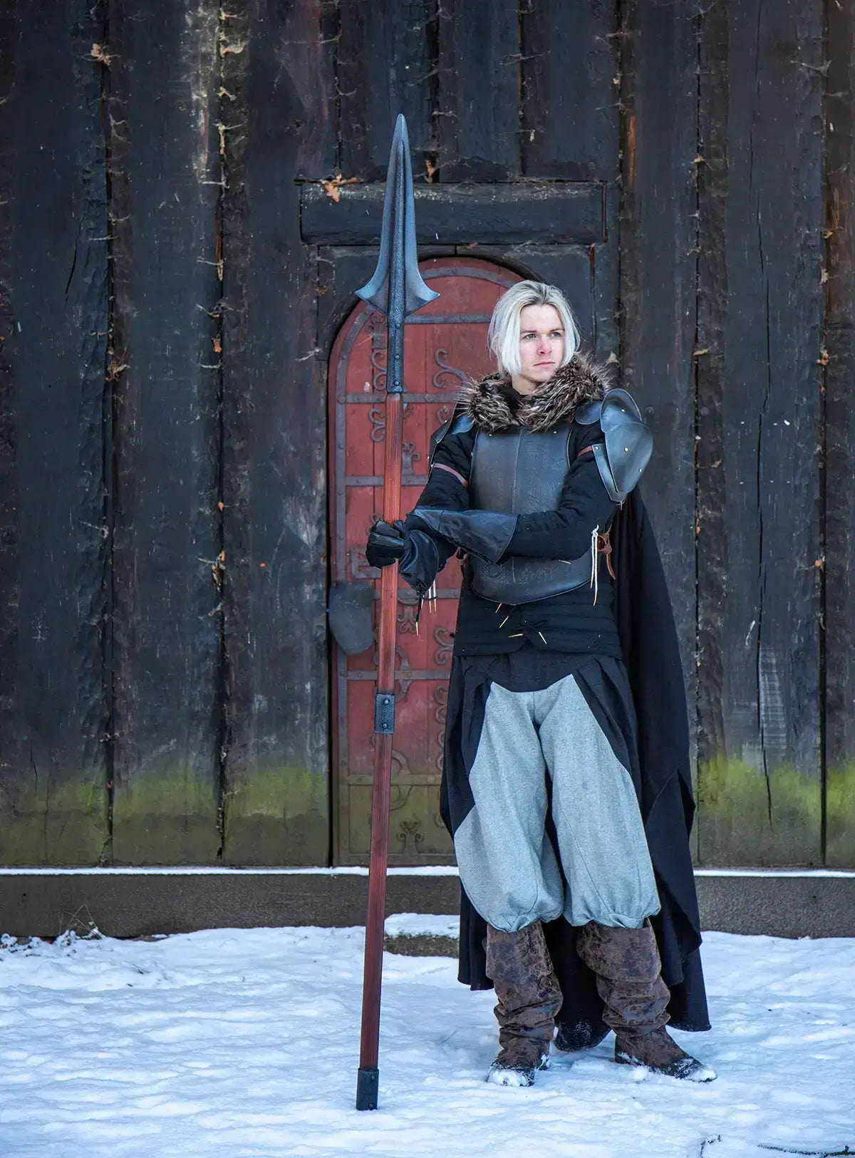 A warrior in a winter setting, holding a spear, dressed in armor and a cloak, against a wooden background.