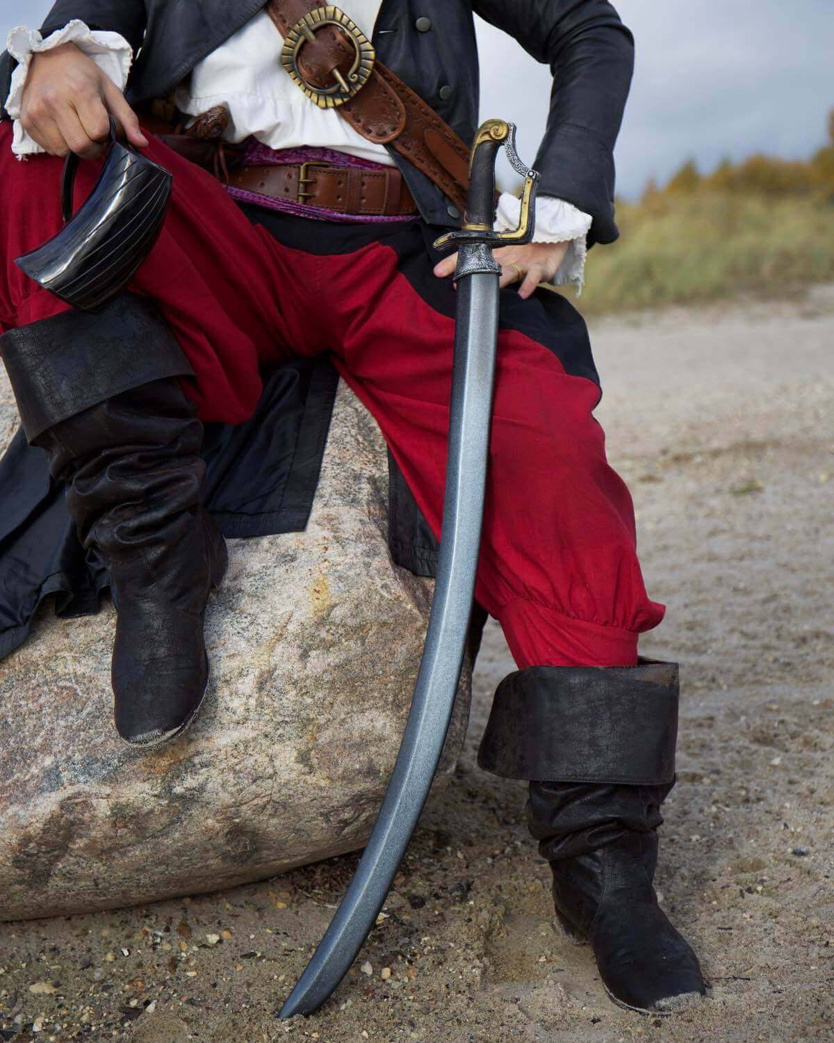 A pirate in red pants and black boots, resting a sword on a rock, showcasing a rugged outdoor setting.