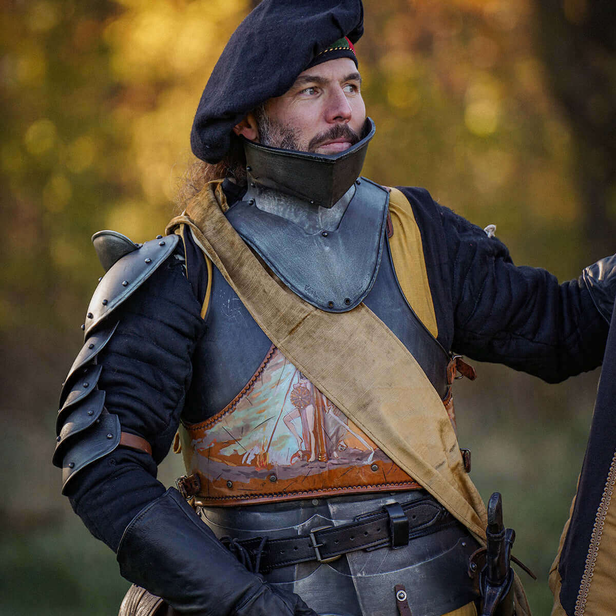A medieval knight wearing armor and a beret, posed in a natural outdoor setting with autumn foliage.