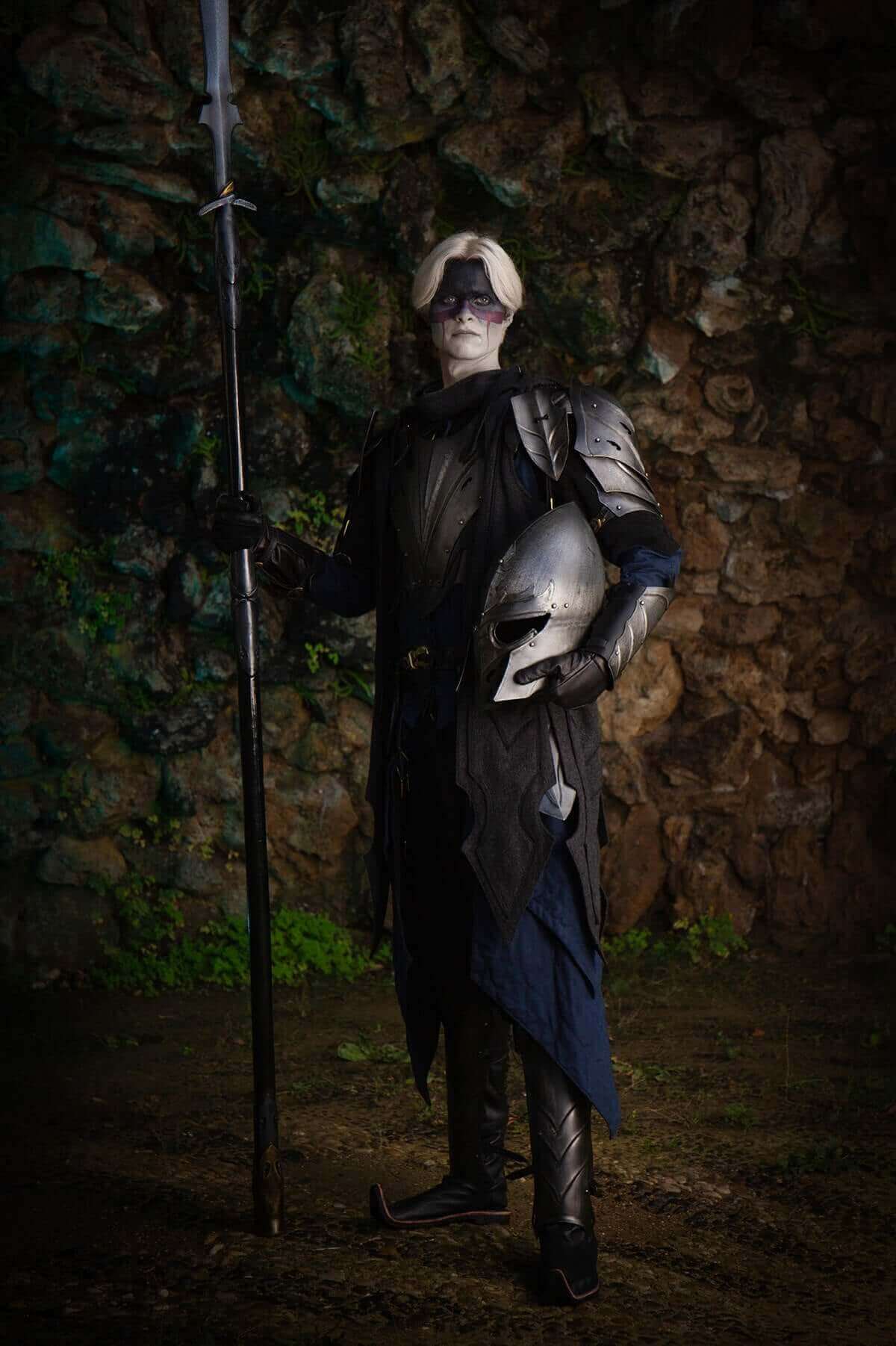 Person in dark costume holding a sword and helmet against a textured wall.