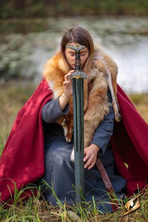 Woman in a red cloak kneeling with a sword by a lake, embodying a fierce warrior spirit.
