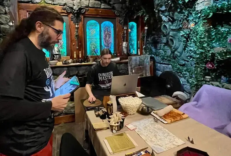 Two gamers in a fantasy-themed room preparing for a tabletop game, with snacks and game materials on the table.