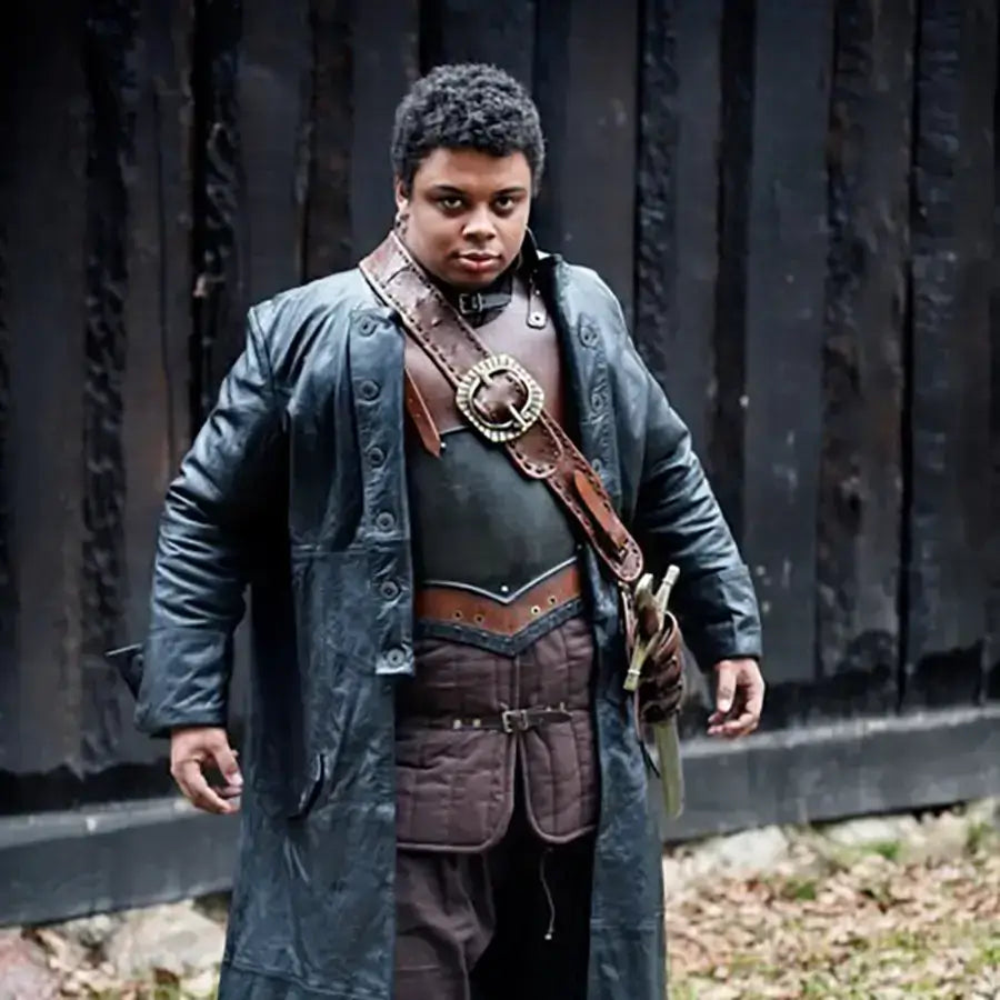 Person in a dark coat with a belt, standing confidently against a rustic wooden backdrop.