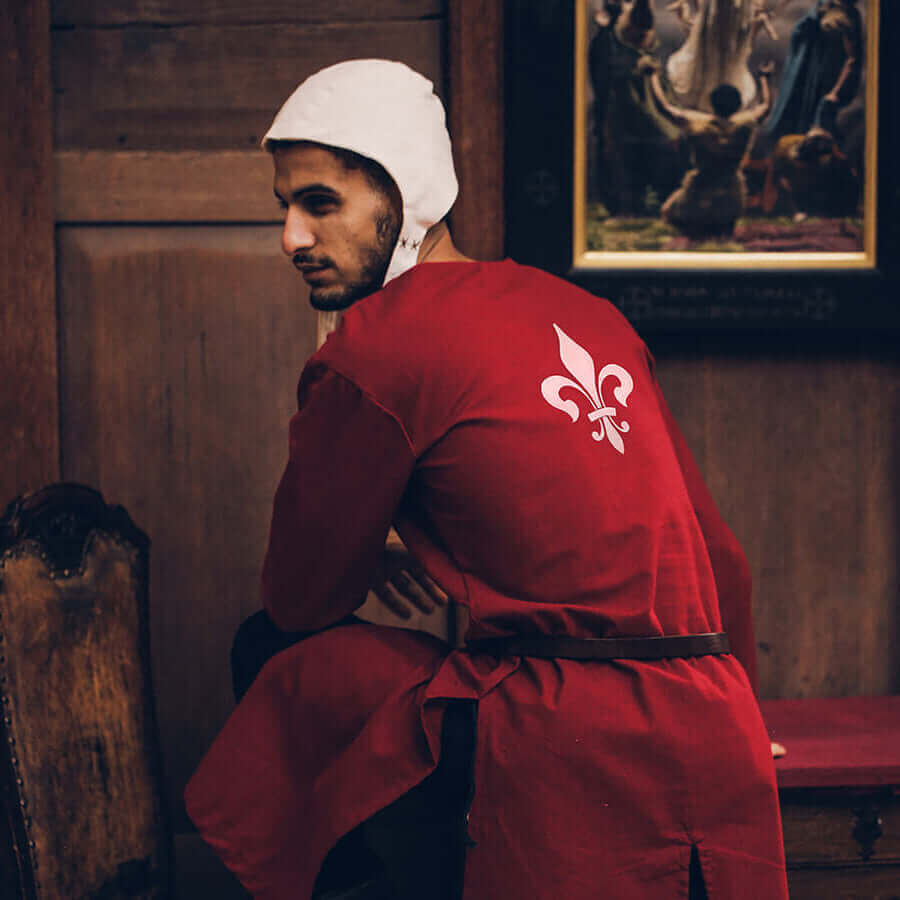 Person in red outfit with white emblem standing in a room with wooden walls and a painting.