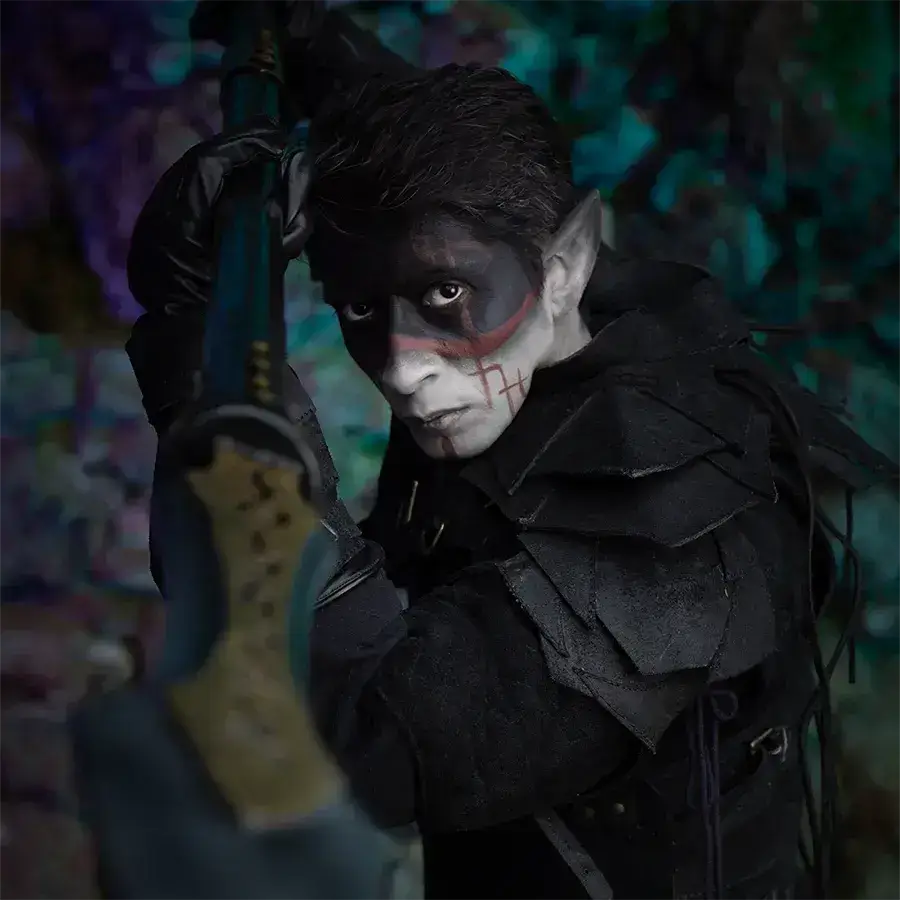 Person in a dark fantasy costume with makeup, holding a sword in a mysterious, colorful forest setting.