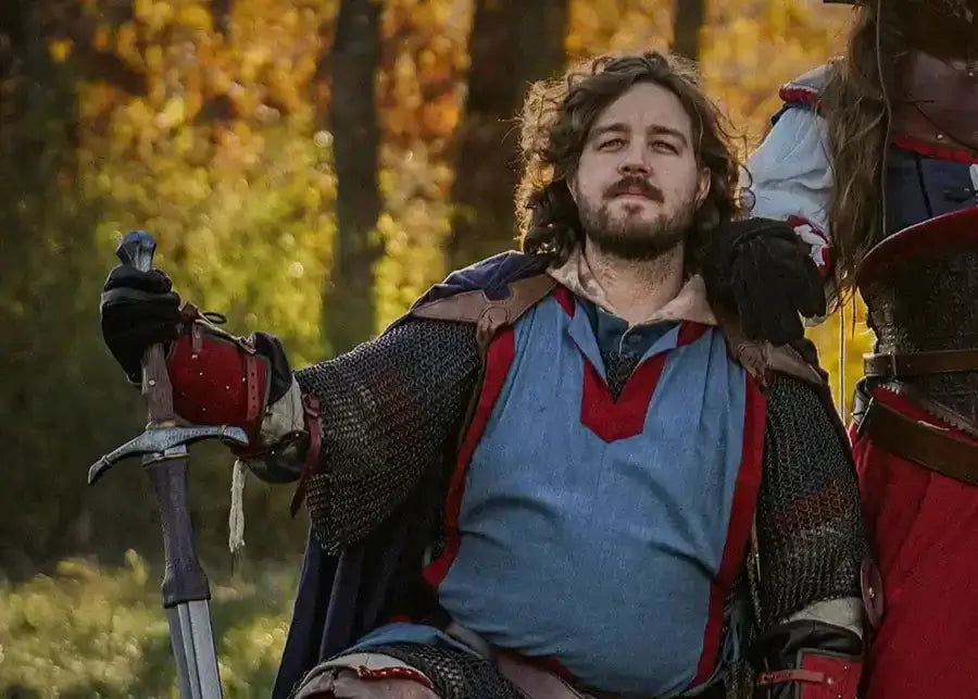 LARP participant in chainmail and a blue tunic resting with a hand on a sword, with another costumed figure standing beside him.