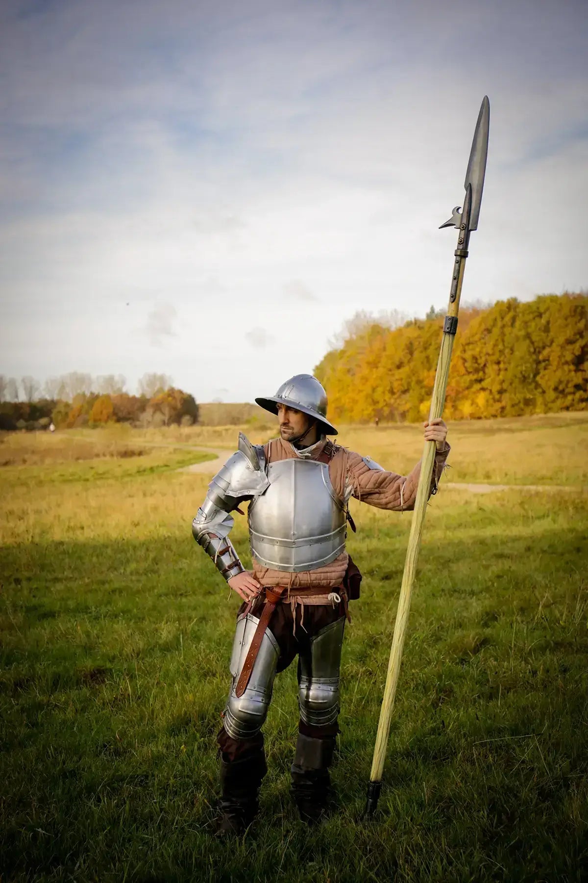 A historical reenactor in armor holding a spear stands in a scenic autumn landscape with trees.