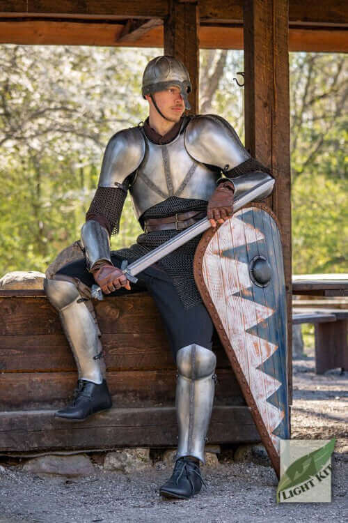 Knight in medieval armor sitting with a sword and shield, showcasing historical costume elements in a natural setting.