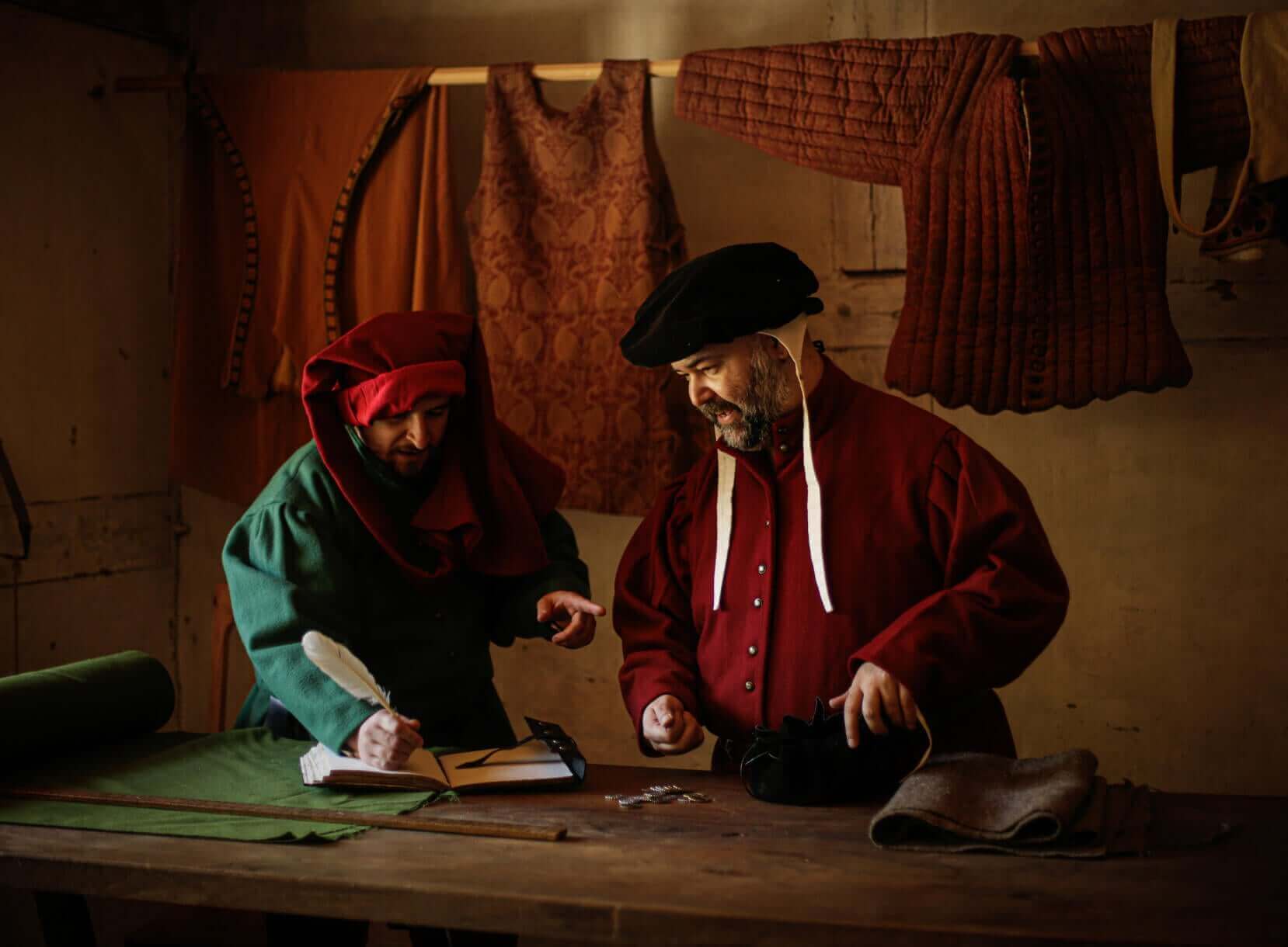 Two historical craftsmen in period attire working at a wooden table with fabrics and tools in a rustic workshop.