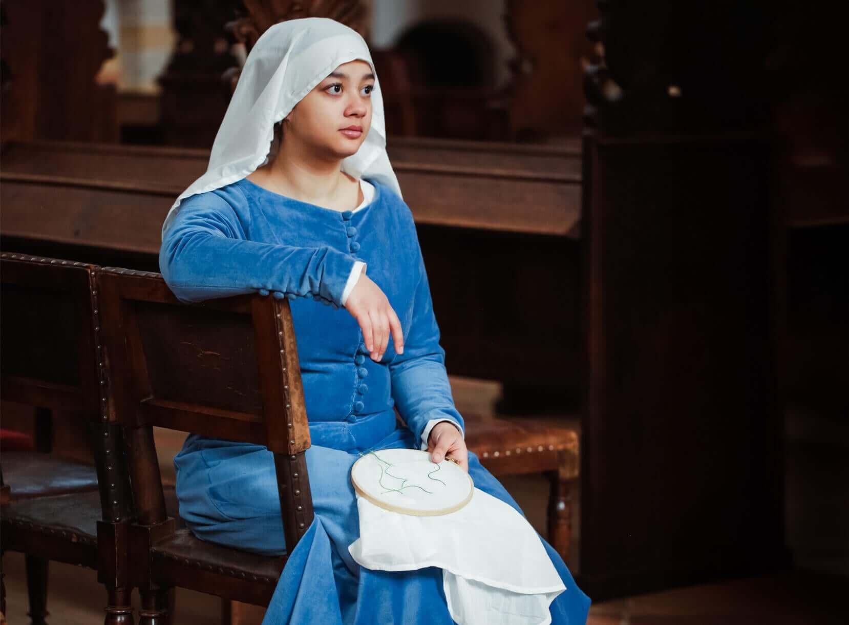 Woman in blue late medieval dress and white headscarf, holding an embroidery hoop.