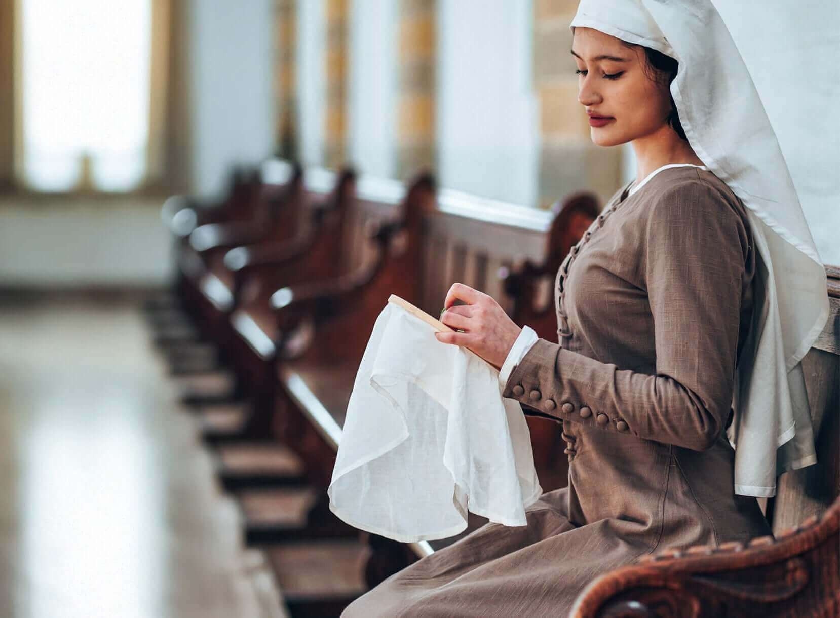 Woman in medieval clothing sewing in a church pew.