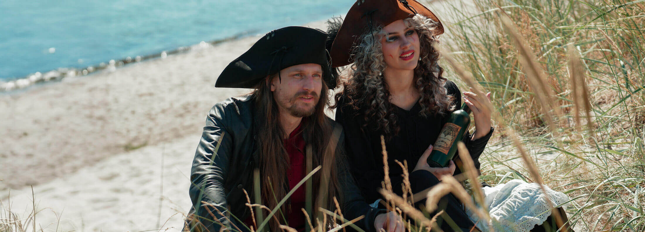 A pair of pirates sitting on the beach, dressed in costumes and sipping from a bottle.